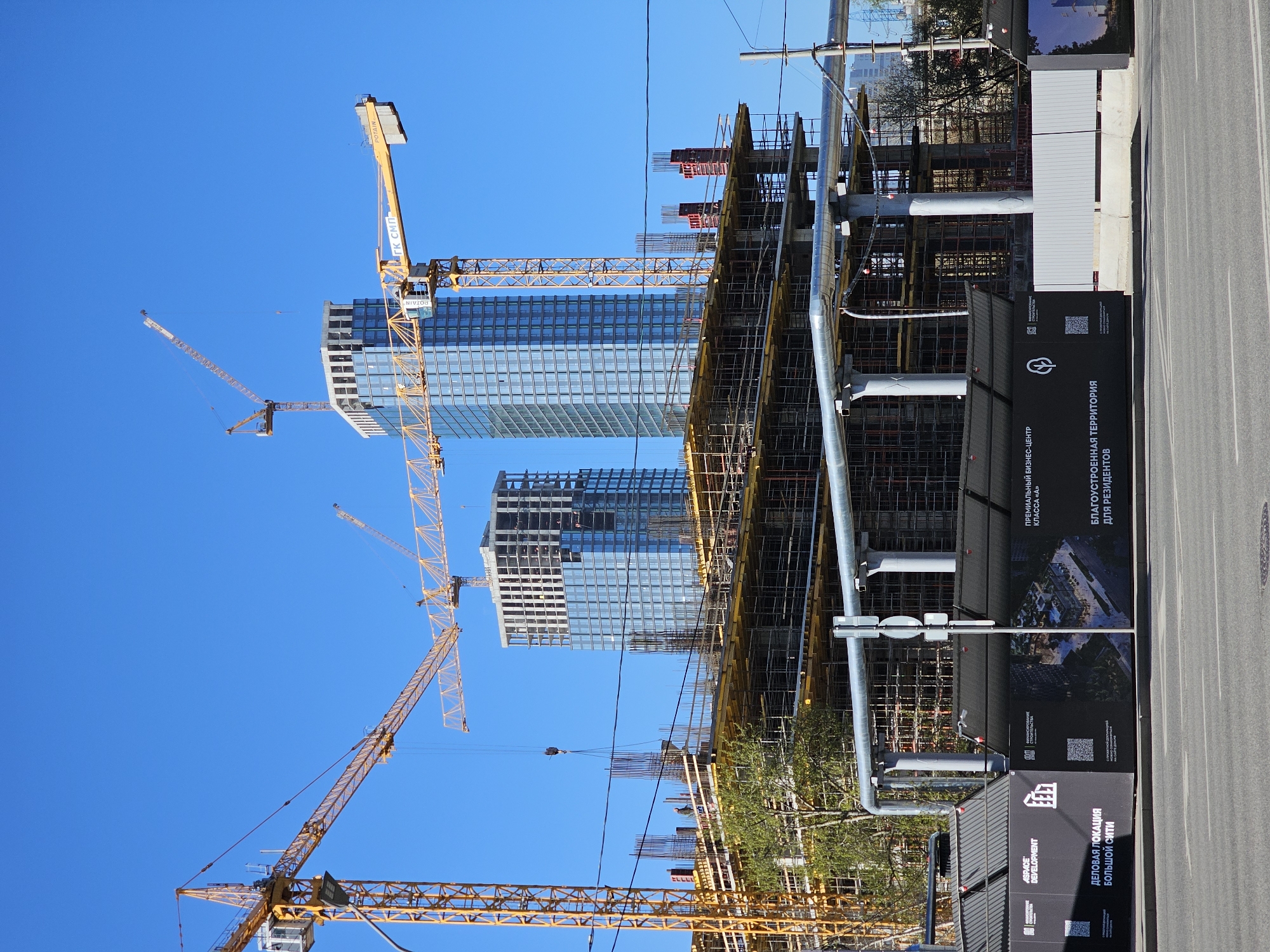 The image shows a construction site in Moscow, Russia, with several high-rise buildings under construction. There are cranes in the foreground and background, and scaffolding surrounds the buildings. Large advertisement boards are posted near the road. The sky is blue and clear.