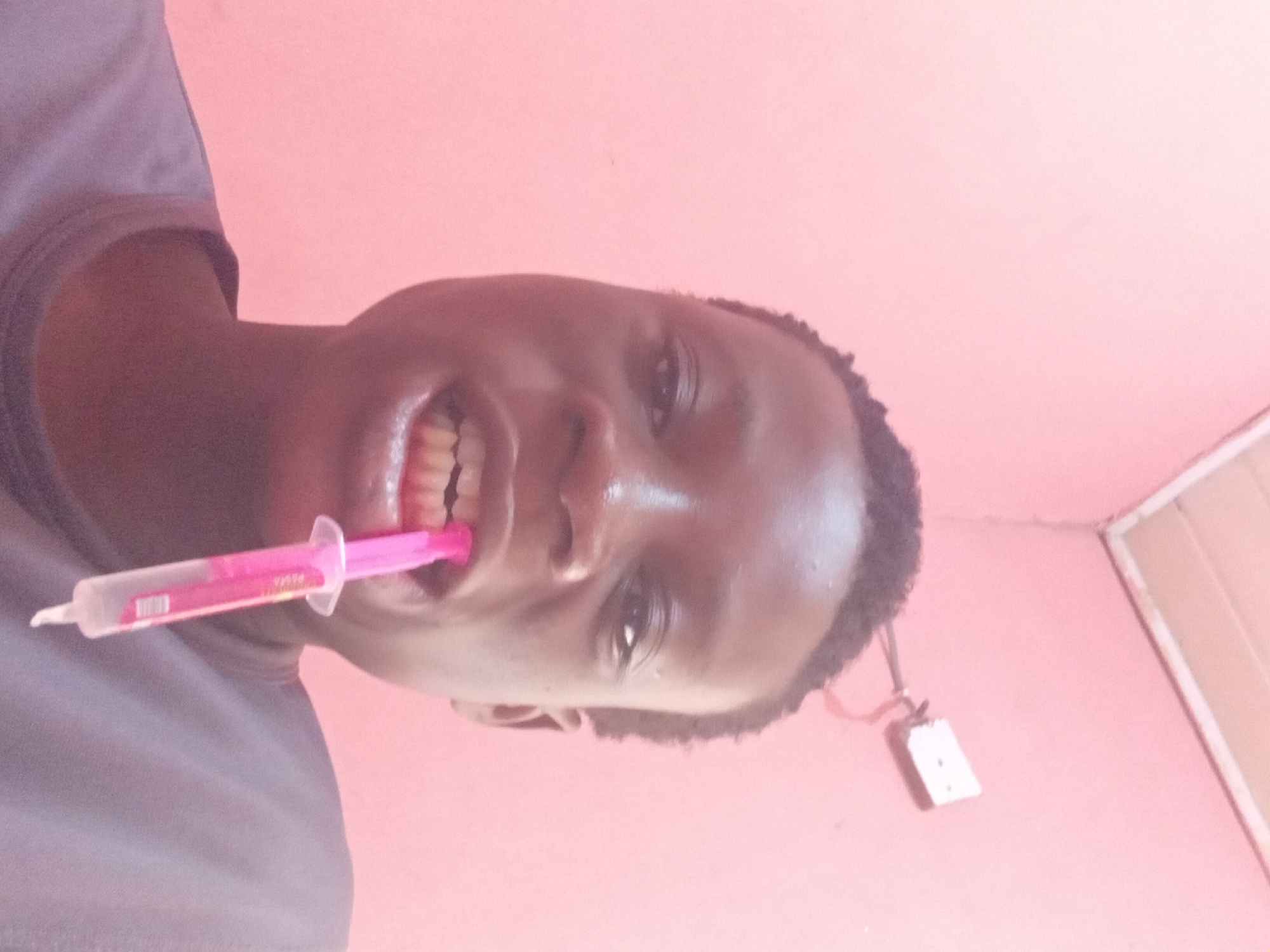 A close-up, low-angle selfie shows a young person of African descent with dark hair and dark eyes. They are wearing a grey t-shirt and have their mouth open, holding a pink syringe-like object in their teeth. The background is a solid, light pink wall with a white electrical outlet visible on the upper right. The lighting is bright, creating some glare on the person's forehead and cheeks. The overall impression is of an informal, indoor setting.