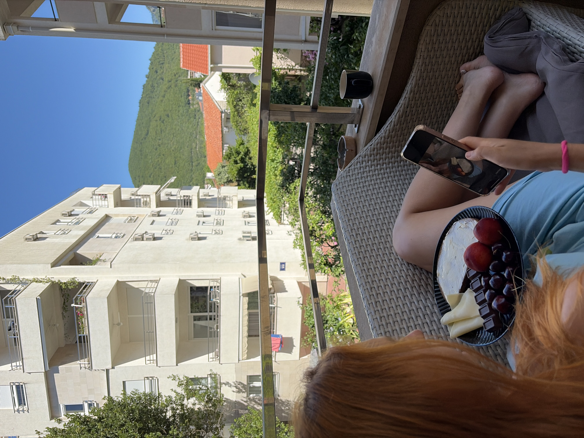 A woman with red hair sits on a wicker chair on a balcony, holding a smartphone in her right hand. She is looking at the screen, which displays a photo of food. In her lap is a dark plate with a breakfast of cheese, chocolate, cherries, and what appears to be buttered toast or a pastry. A dark mug and an ashtray are on the ledge of the balcony. In the background, a white apartment building with balconies and air conditioning units is visible, as well as green trees and a distant green mountain covered in foliage, characteristic of the city of Budva, Montenegro. The sky is a clear blue.