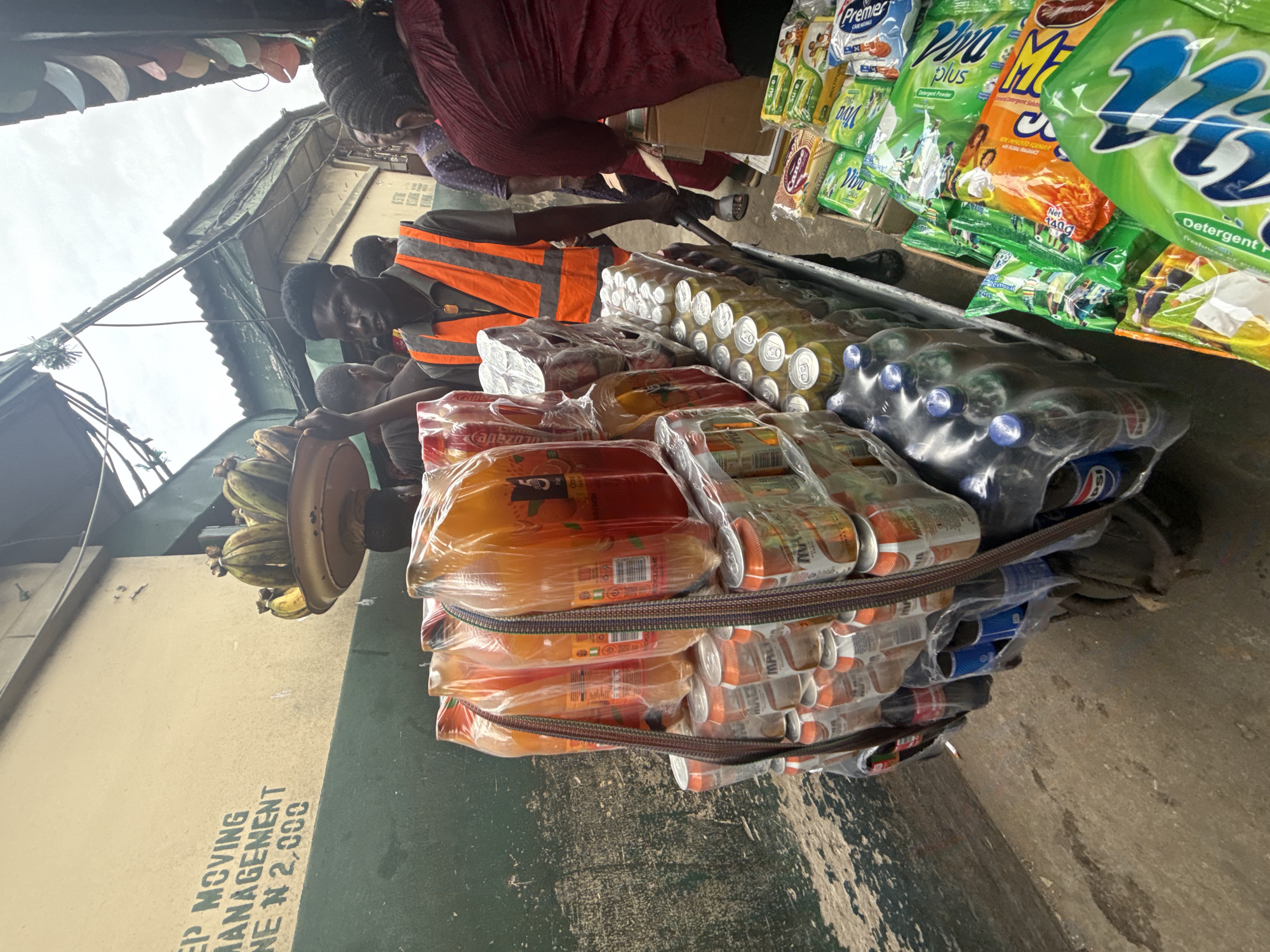 In a Lagos, Nigeria market, a vendor pushes a handcart loaded with assorted beverages and packaged goods. The vendor, a young Black man, wears an orange high-visibility vest over a grey shirt and appears to be focused on navigating the crowded pathway. A bunch of bananas rests on a stand atop the handcart. The cart is laden with stacks of bottled drinks, including large orange bottles and smaller cans of what appears to be malted beverages. Several plastic-wrapped cases of bottled water are also visible, some bearing Pepsi branding. In the foreground and to the right, an assortment of detergent and other packaged cleaning supplies is displayed. The market environment is characterized by narrow walkways, overhead wires, and corrugated metal roofing, common in such urban commercial settings. The wall on the left side of the image has faded green and yellow paint with some faded text that appears to be in English, possibly a business or management instruction. The ground is dirt and concrete, showing signs of wear and tear from constant foot traffic.