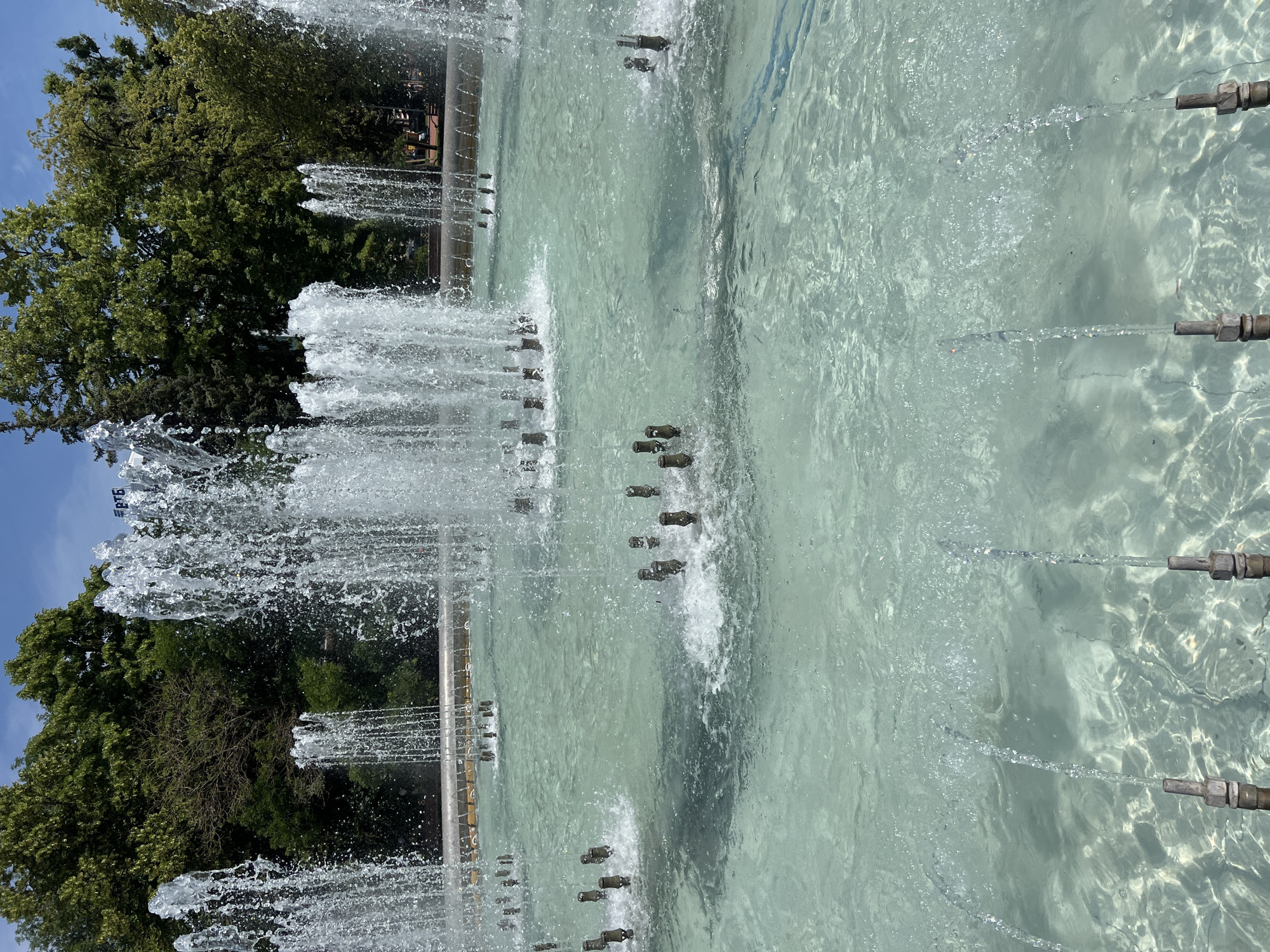 This image depicts a large public fountain in the city of Krasnodar, Russia. The fountain is active, with numerous jets of water spraying upwards in various patterns, creating a dynamic and refreshing scene. The water in the fountain basin is a clear, light turquoise color, and sunlight reflects off its surface, casting shimmering patterns. Several metallic nozzles are visible, protruding from the bottom of the basin, from which the water is being propelled. In the background, a dense line of lush green trees forms a natural backdrop, suggesting a park or garden setting. The sky above is a clear blue with a few wispy clouds. The overall impression is one of a serene and pleasant outdoor environment, characteristic of a public space designed for relaxation and enjoyment. The prominent "РТБ" logo visible on the fountain structure indicates it is located in or is sponsored by the R.T.B. company.