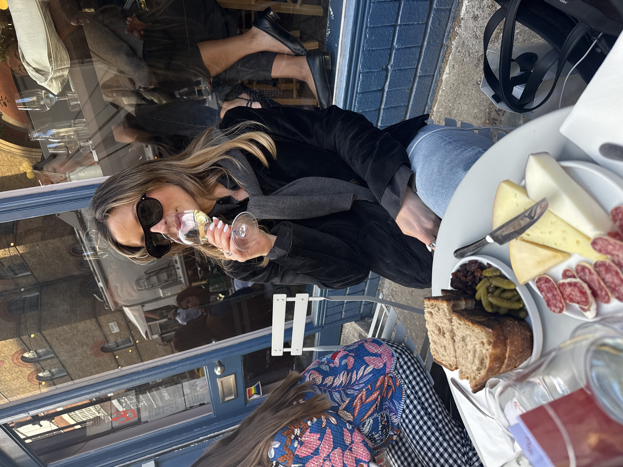 A sunny outdoor scene captures two individuals enjoying a meal and drinks at an urban cafe or restaurant. In the foreground, a woman with long, light brown hair, wearing sunglasses, a black jacket, and light blue jeans, is seated at a white table. She holds a wine glass with a light-colored beverage, taking a sip. Her nails are painted white.
Opposite her, a second person is partially visible, showing their lap and lower torso, clad in a blue patterned top and a black and white checkered skirt or dress.
On the white table between them rests a platter featuring a charcuterie and cheese board. It includes various slices of cheese, sliced salami or cured sausage, gherkins, what appears to be dried fruit, and several slices of rustic bread, alongside a knife. Other clear glasses and bottles are visible, primarily as reflections.
The background reveals a dark blue storefront or building facade. A large window in this facade heavily reflects the surroundings, showing an urban environment with brick buildings, some featuring arched windows, suggesting a street view. The reflections also show parts of the establishment's interior, with bottles and glasses. A small rainbow-colored flag is discreetly visible on the blue facade.
The bright natural light indicates it is daytime, and the relaxed postures suggest a pleasant, leisurely outing. A black tote bag is seen next to the prominent woman's chair. The overall setting is consistent with an outdoor dining area in a city like London, United Kingdom.