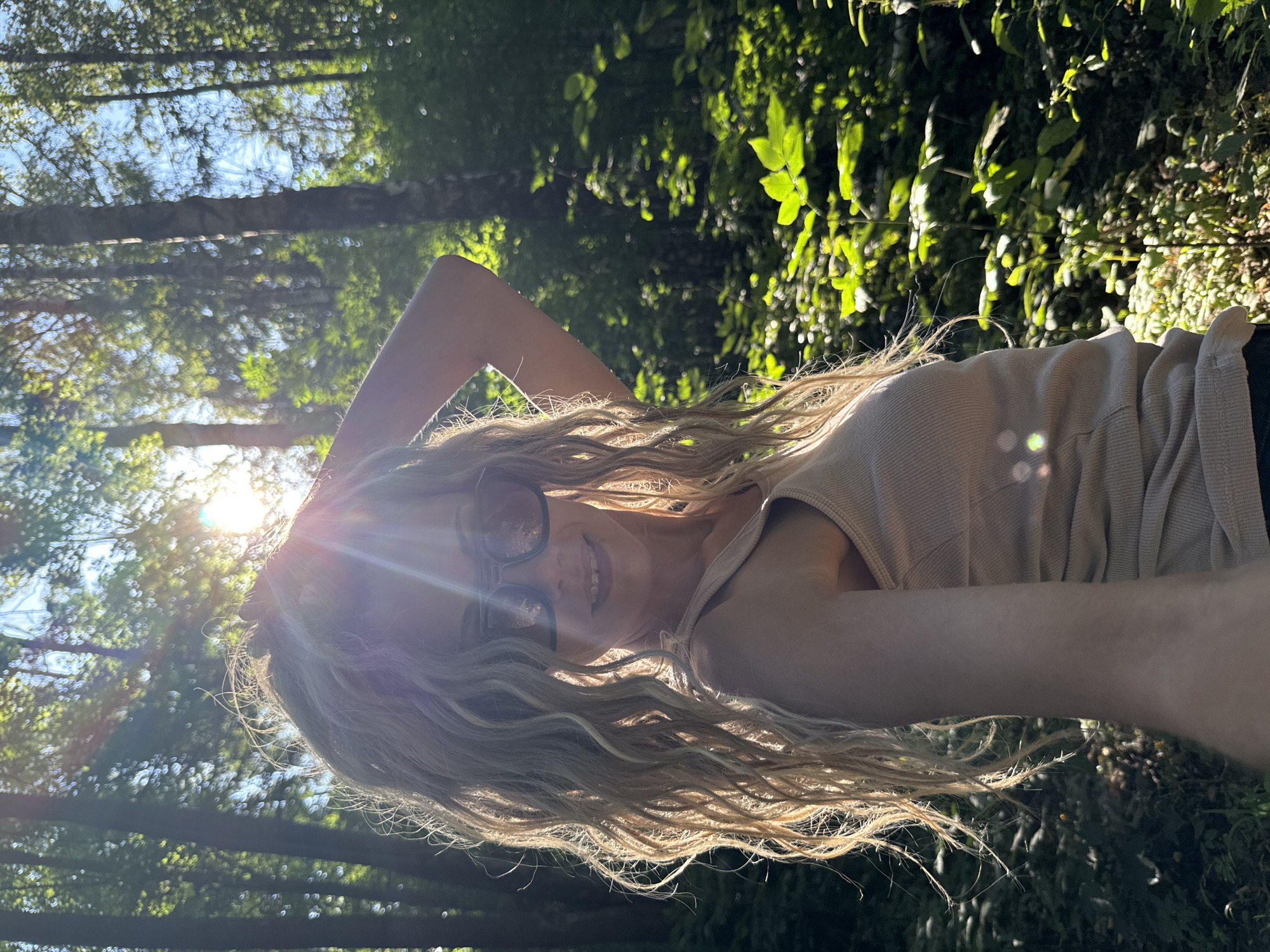 The media file is a selfie taken outdoors in a wooded area. A young woman with long, wavy blonde hair and sunglasses is visible. She is wearing a light-colored sleeveless top. The sun is shining brightly, casting lens flares and illuminating her hair and face. She has her arm raised, with her hand behind her head. Her expression appears relaxed and happy. The background consists of trees and foliage, with sunlight filtering through the leaves, suggesting it is daytime, likely morning or afternoon given the quality of light. The overall scene evokes a sense of a warm, sunny day spent in nature.