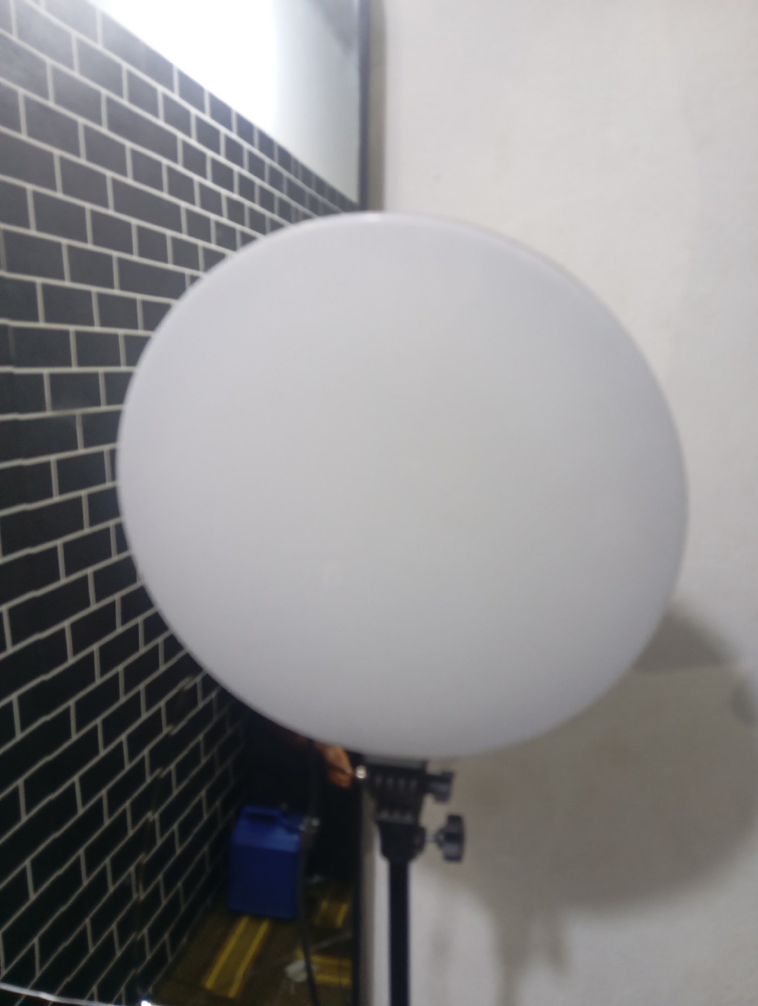 A softbox, a piece of lighting equipment used in photography, is the primary subject of this image. It is a large, white, spherical diffusion dome mounted on a black stand with several adjustment knobs. The softbox is positioned in the foreground, partially obscuring the background. To the left of the softbox is a wall covered in black brick-like tiles with white grout. In the lower-left corner of the background, a bright blue container or box is visible on the floor, along with some yellow markings on the floor. The rest of the background consists of a plain white wall and some dark areas, suggesting an indoor setting. The image quality is somewhat blurry, making it difficult to discern fine details. There is no discernible location in Jauro Yinu, Nigeria, evident from the visual information provided in the image.