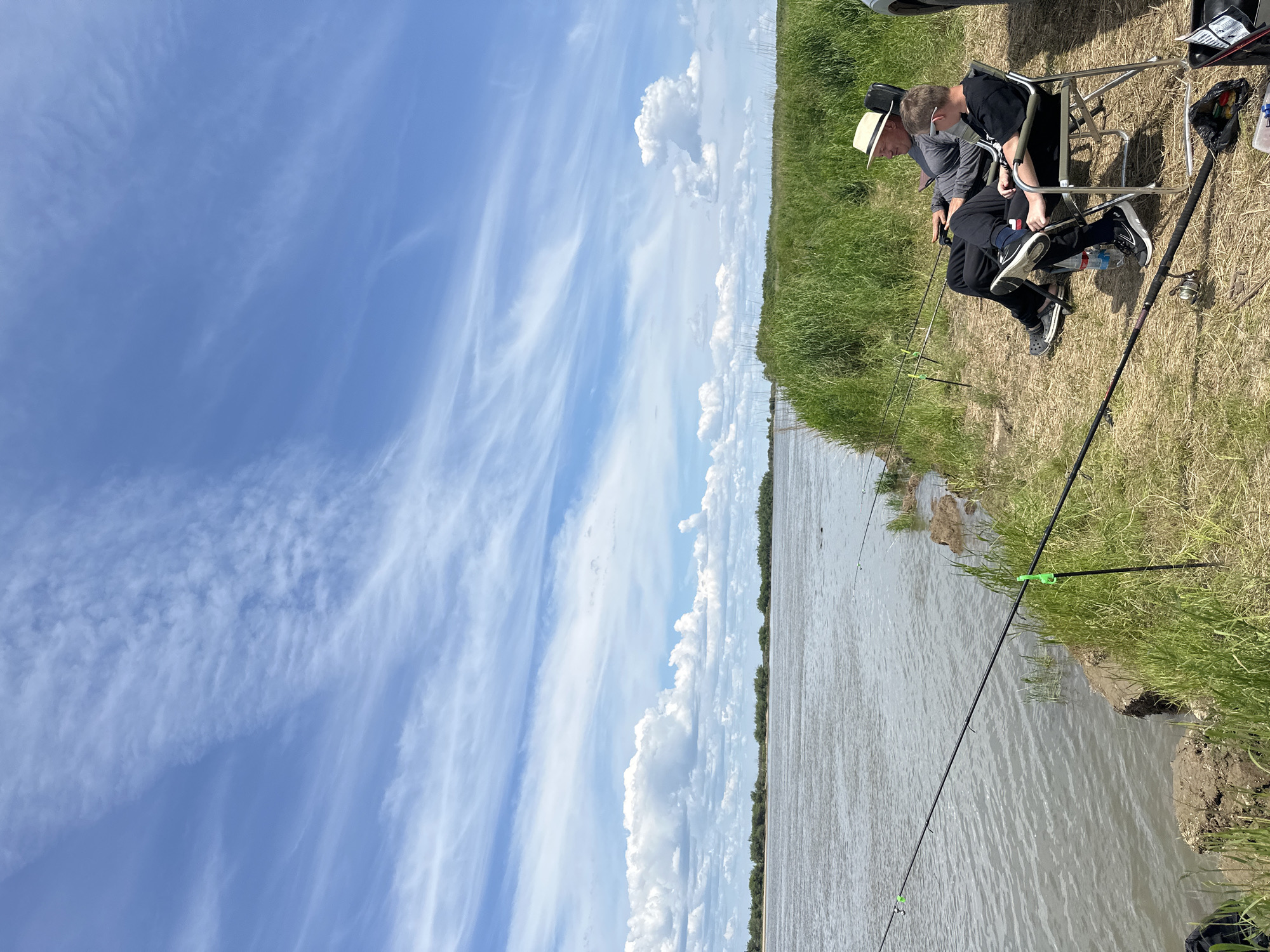 Two individuals are seated in camping chairs on a grassy embankment beside a wide, calm body of water, possibly a river or lake. The person in the foreground, a younger individual wearing a black t-shirt and black pants, is looking towards the water. The person behind them, an older individual wearing a grey long-sleeved shirt and black pants, is wearing a light-colored straw hat and sunglasses, and appears to be looking at a mobile phone. Both individuals have fishing rods set up in front of them, with lines extending into the water. The environment is rural, with tall green grass and reeds lining the bank. The sky is partly cloudy with dramatic, wispy white clouds against a blue expanse. The overall impression is a serene outdoor scene, likely a fishing trip. The image does not provide any specific visual cues to determine its location within Im. Tel'mana, Russia.