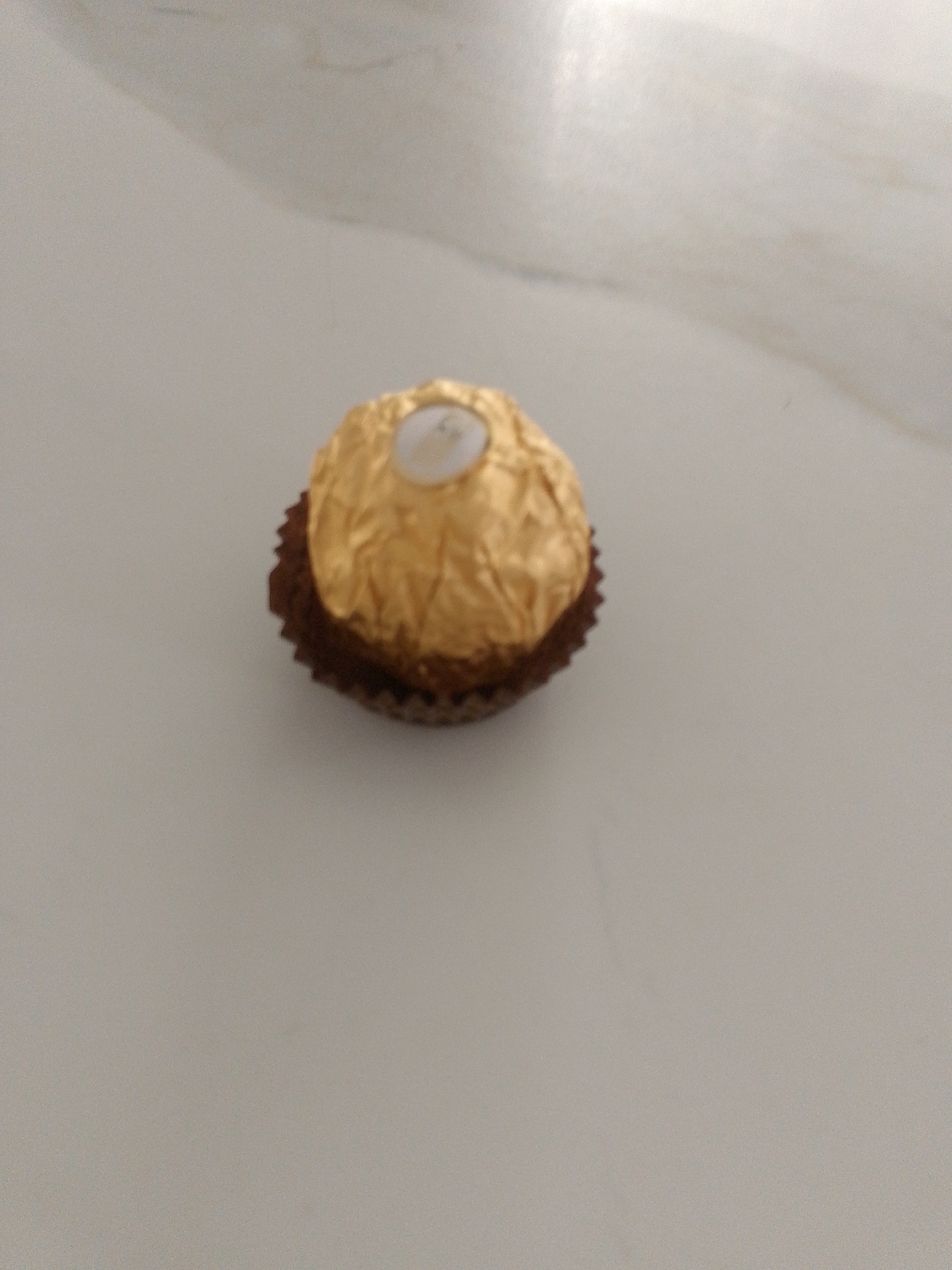 A single Ferrero Rocher chocolate is centered on a pale, speckled surface, likely a countertop or table. The chocolate is wrapped in its signature gold foil, which is crinkled and shiny, reflecting the light. A small, white, circular embellishment is visible at the top of the foil. The bottom of the chocolate is encased in a brown, ruffled paper liner. The background is softly blurred, indicating a shallow depth of field, and appears to be a neutral, light-colored material with subtle variations in tone and texture.
