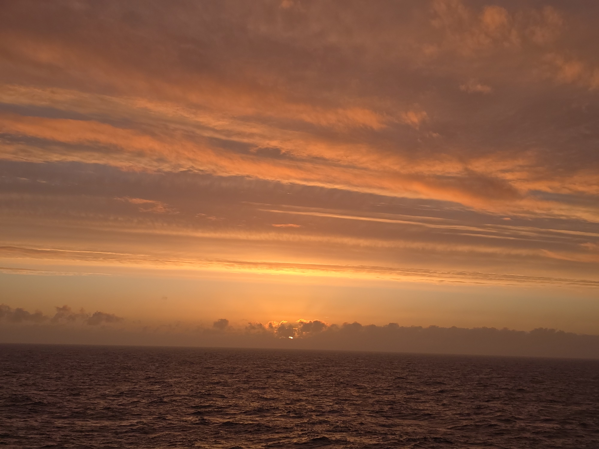 The image depicts a sunset over the ocean. The sky is filled with orange and pink clouds. The sun is partially obscured by a layer of clouds near the horizon. The sea reflects the colors of the sky. The location is unspecified.