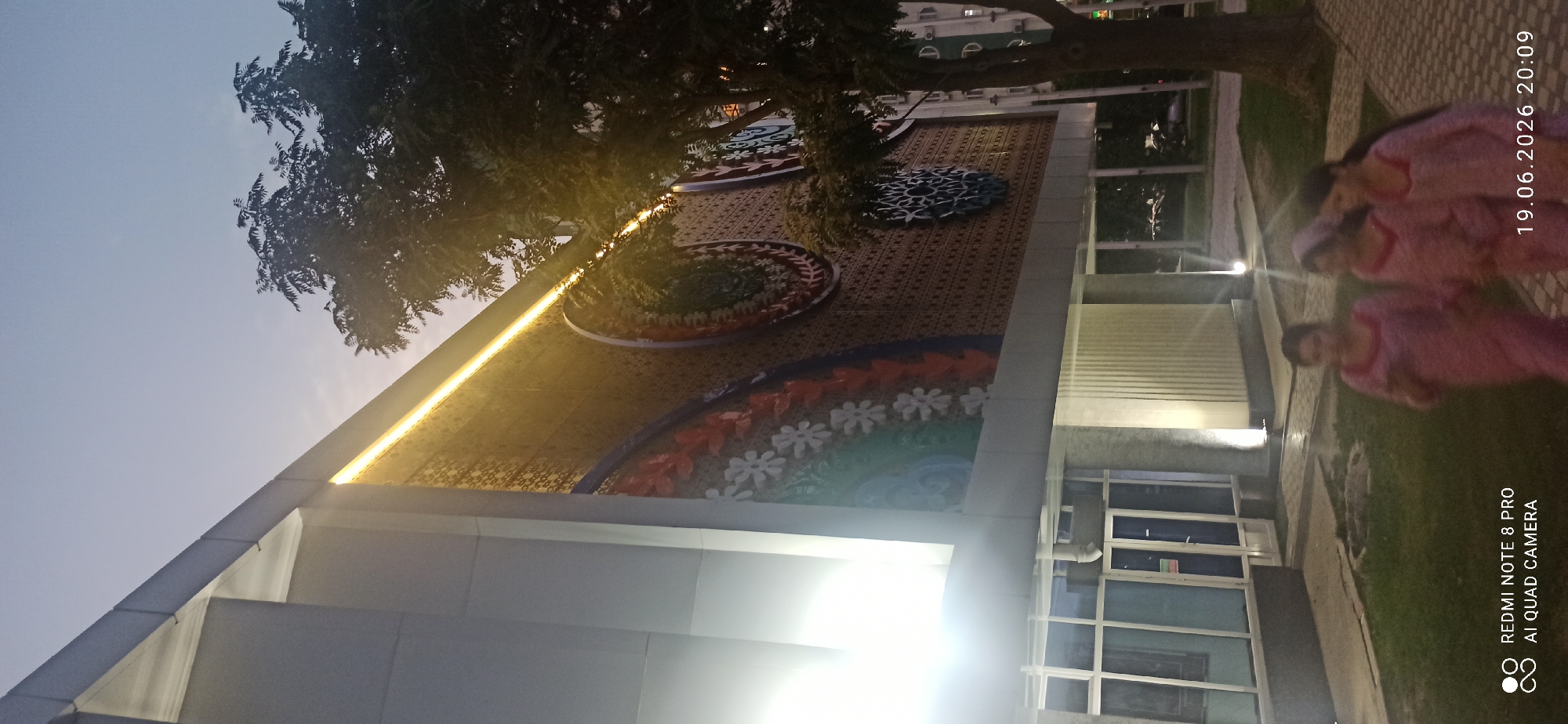 This media file shows an outdoor scene with a building facade decorated with colorful circular motifs. A tree with dark foliage is partially obscuring the building and the sky. The sky appears to be twilight or dusk, with a soft, muted light. A strip of bright yellow light runs along the edge of the building, illuminating the artwork.
In the foreground, to the right of the building, three people are visible, out of focus. They are wearing pink clothing. The lighting in this area is dim, and the overall mood is relaxed. The ground is paved with stone tiles, and there is grass and some greenery.
Visible text in the bottom right corner reads "REDMI NOTE 8 PRO AI QUAD CAMERA" and a timestamp "19.06.2026 20:09". The timestamp suggests the image was taken in the evening. The scene appears to be in an urban or semi-urban setting, possibly a park or a public space adjacent to a building. The artwork on the building suggests a decorative or artistic purpose. The overall impression is of a peaceful evening captured outdoors.