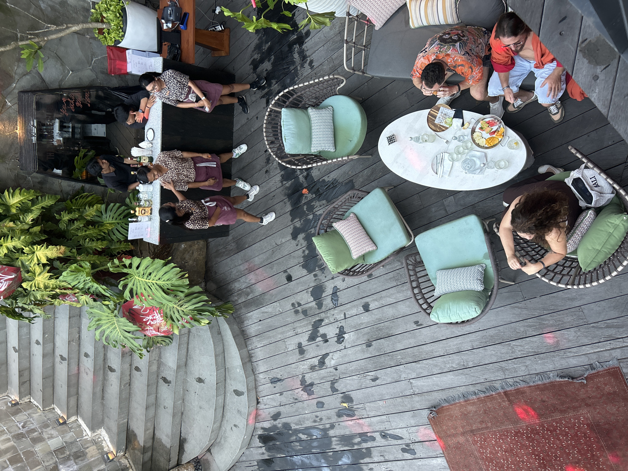 An elevated view captures an outdoor seating area with a bar and several patrons in Sukawati, Indonesia. The scene features a grey stone staircase on the left, leading down to a weathered wooden deck. Lush green foliage with large leaves and red accents is prominent, particularly near the staircase and bar area.
A dark-colored bar counter is staffed by four individuals wearing matching patterned shirts and maroon aprons. They appear to be interacting with each other and serving guests. Behind the bar, a glass-fronted cabinet displays bottles.
Several seating areas are arranged on the deck. One area features two light-colored, modern armchairs with light green cushions and decorative pillows. Another section includes a low, oval-shaped white table with glasses, bottles, and a fruit platter. A dark grey sectional sofa with striped cushions is positioned near this table.
Four patrons are seated in this area. Two men are seated on the sofa, one wearing a vibrant orange and black patterned shirt, the other in a red shirt and light blue pants. Both appear to be eating and drinking. A woman with curly brown hair is seated in one of the wicker armchairs, wearing a dark top and brown pants, with a white tote bag beside her. Another person, partially visible, is seated in the other armchair.
The wooden deck has marks and stains, suggesting recent activity or weathering. A red patterned rug with fringe lies on the deck to the left of the seating area. The overall impression is a lively outdoor dining or bar environment.