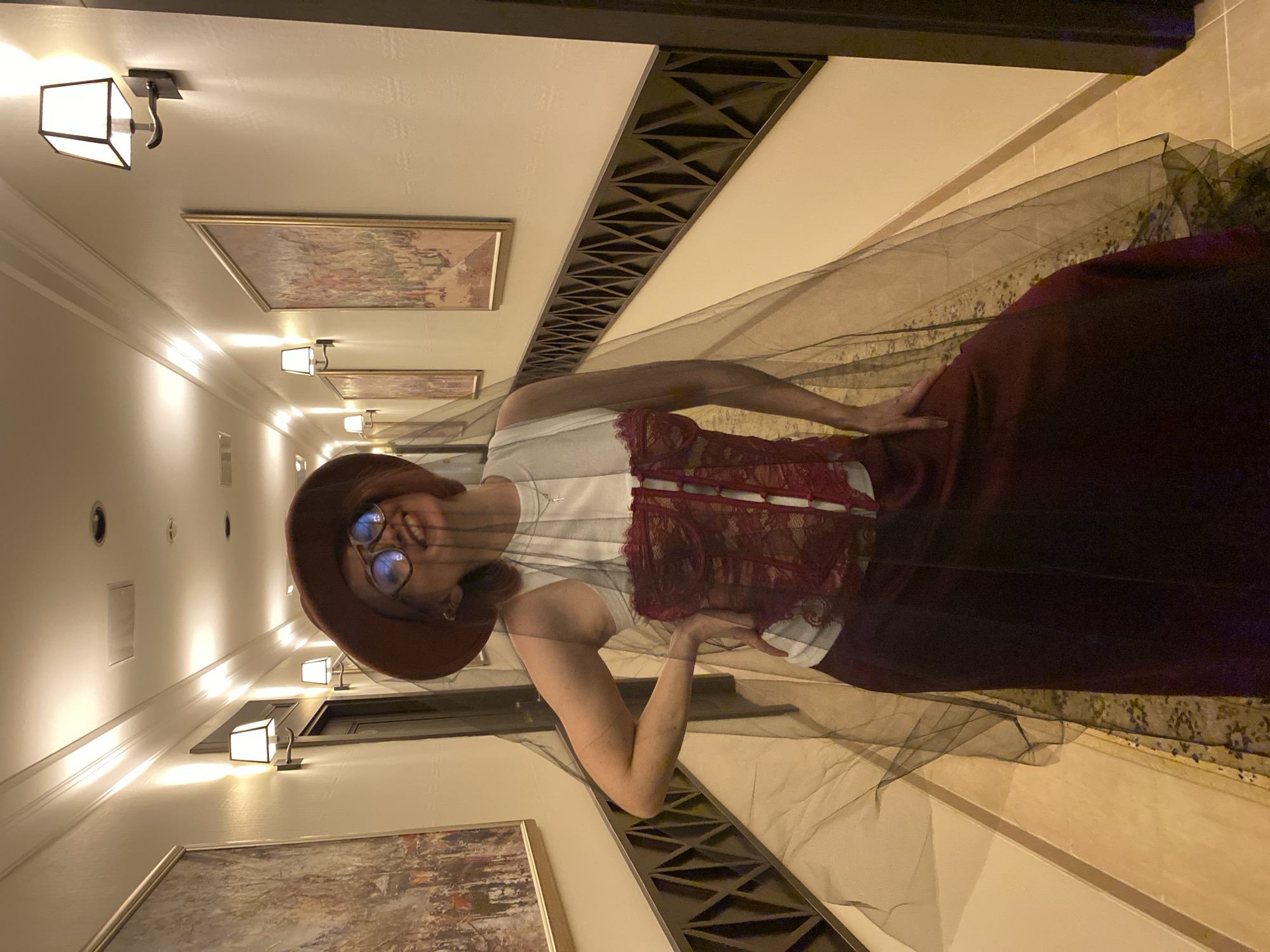 A woman wearing glasses and a hat poses in a hallway. She is wearing a white top with burgundy lace and a long, dark skirt, draped with a dark veil. The hallway features artwork on the walls, sconce lighting, and a decorative ceiling element. The location appears to be indoors and is likely not in the city of Yekaterinburg, Russia.