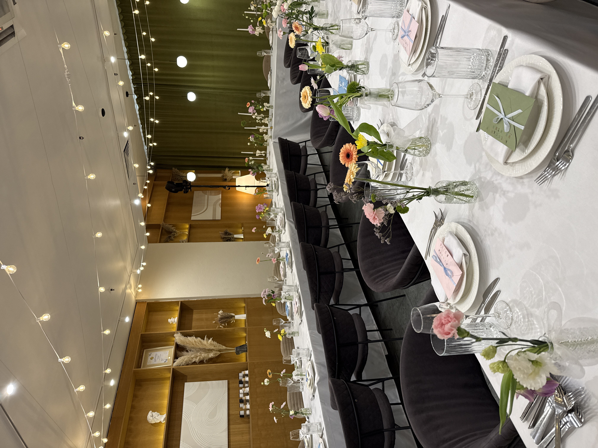 The image shows an interior setting, likely a banquet hall or event space, in Saint Petersburg, Russia, prepared for a formal event.
The dominant feature is a long, white tablecloth-covered table stretching into the distance. It's set with plates, silverware, glassware, and small vases containing single flowers, placed at regular intervals. Each place setting includes an envelope with a ribbon.
Around the table are dark-colored chairs.
In the background, there's a wall with wooden shelving units, which display decorative objects.
Overhead, strings of lights illuminate the scene. There are green curtains to the right of the wooden shelving, and a person standing off in the distance.