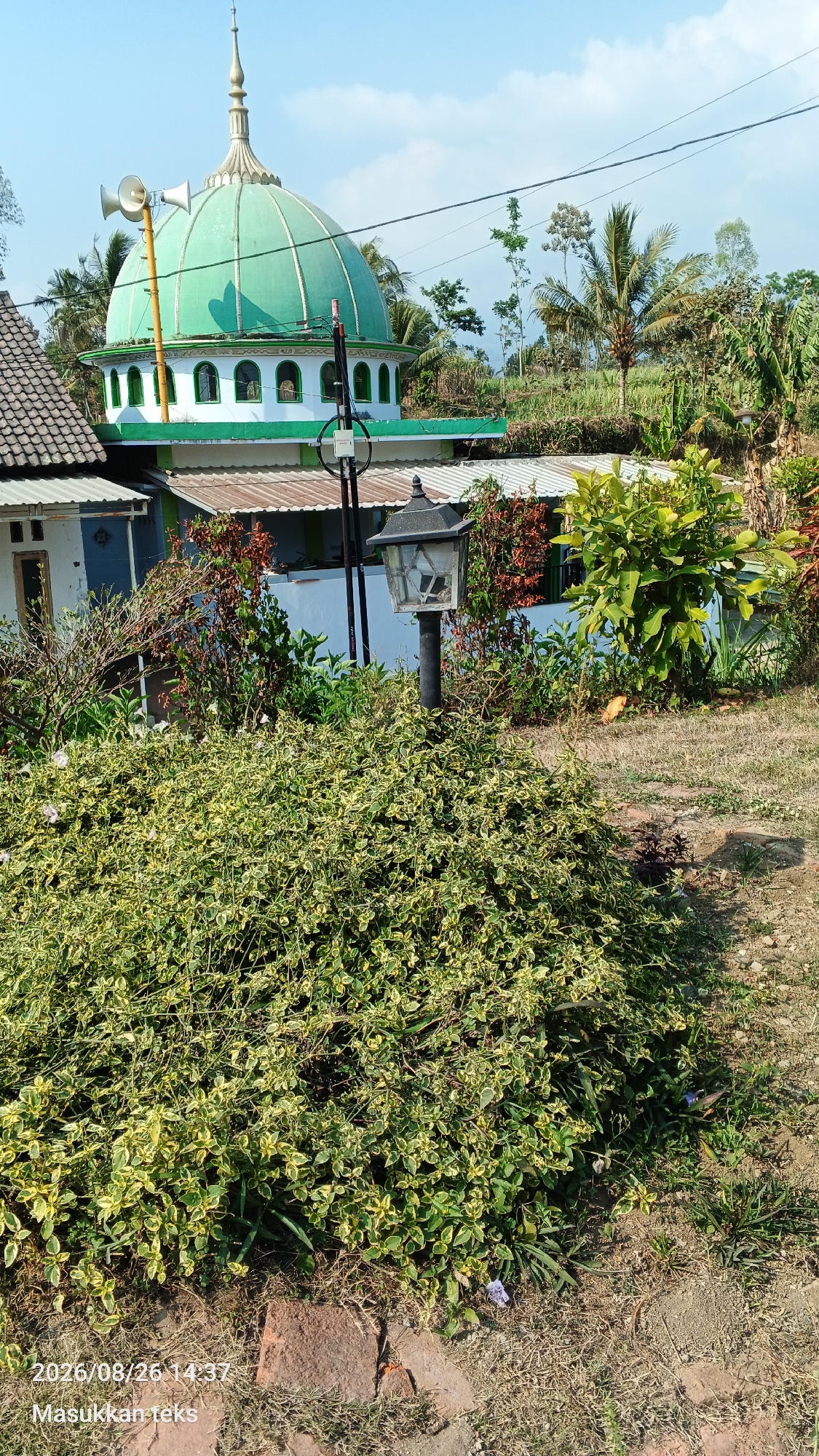 In the city of Jeru, Indonesia, a mosque with a green dome stands tall under a clear blue sky. The mosque features white accents, arched windows, and is topped with a spire. Two loudspeakers are mounted on a pole near the dome. A street lamp with a black base and a broken glass lantern sits in front of a lush green and yellow-variegated bush. Behind the bush, other vegetation and trees are visible, as well as parts of buildings with tiled roofs. The ground is a mix of dry grass and exposed earth. A timestamp "2026/08/26 14:37" and the text "Masukkan teks" are overlaid on the bottom left of the image.