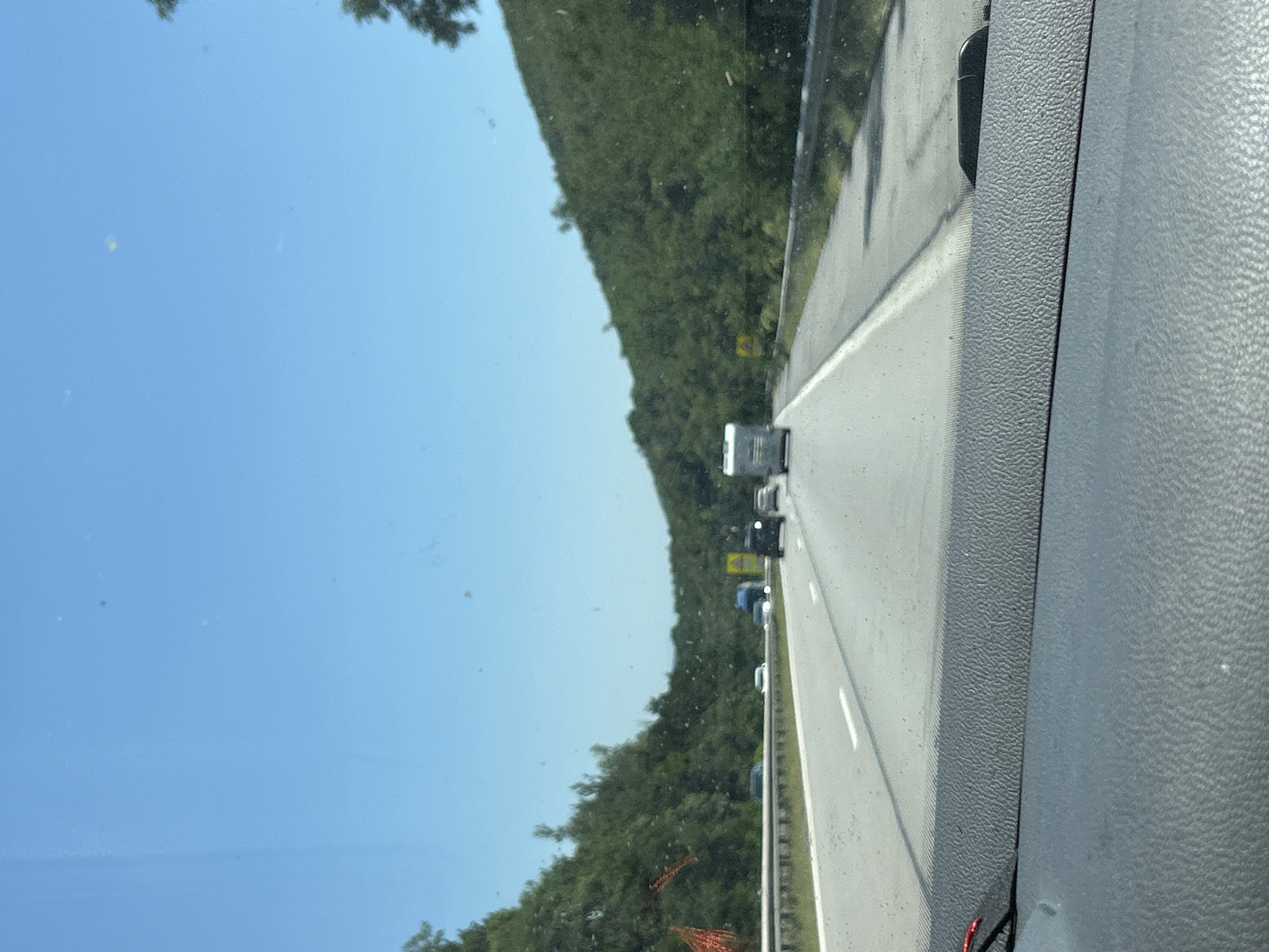 The image depicts a view from inside a vehicle traveling on a highway with clear blue skies overhead. The highway has multiple lanes, and several vehicles, including cars and a camper van, are visible on the road. Dense green foliage covers the hillsides on both sides of the highway, suggesting a natural, possibly mountainous, environment. A metal guardrail runs along the edge of the highway. In the foreground, the textured dashboard of the vehicle is visible. The scene appears to be captured during daylight hours. The location is identified as the city of Dolna Vasilitsa, Bulgaria.
