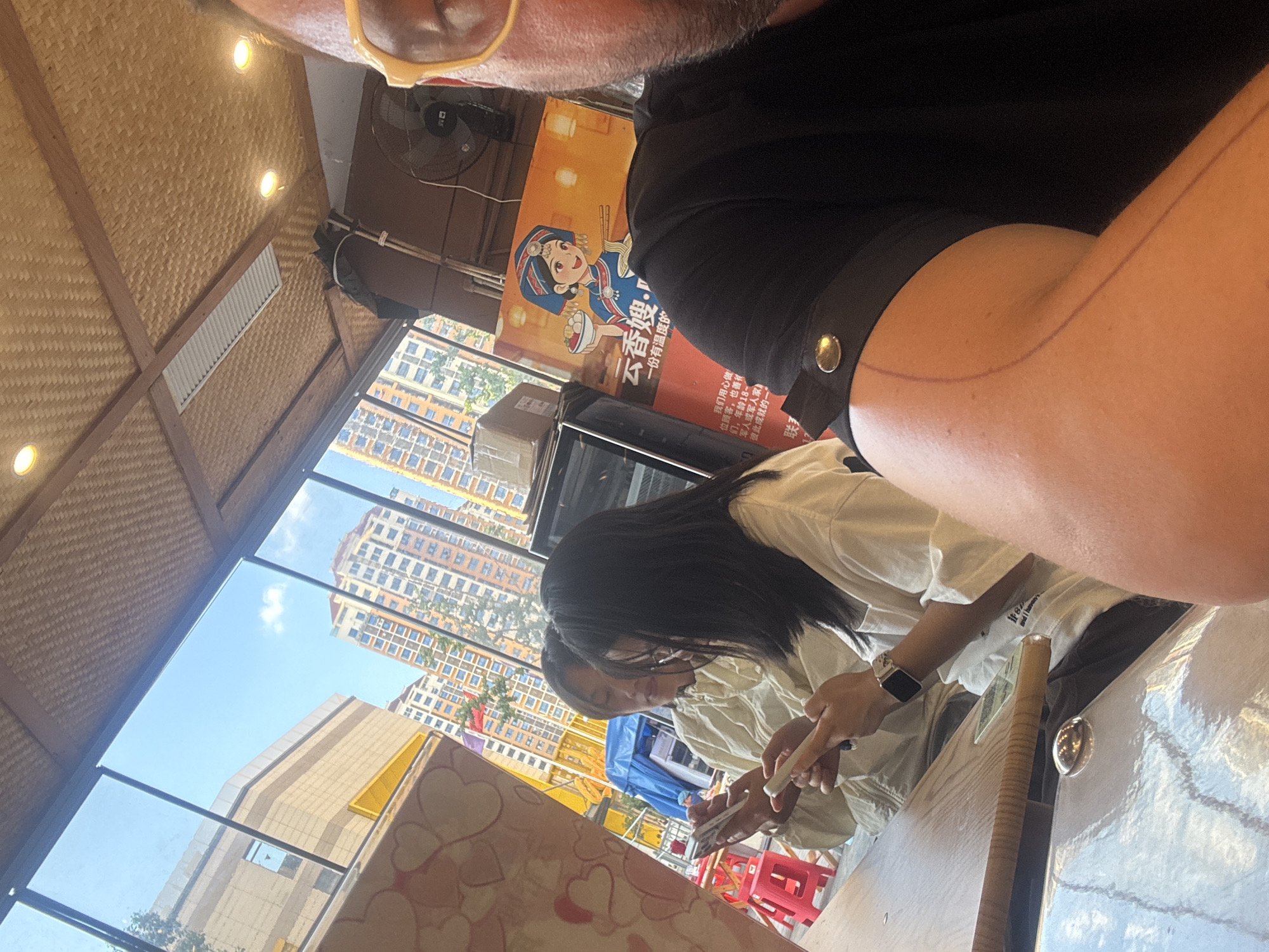 In Suzhou Industrial Park, China, a selfie is taken from the perspective of a person wearing yellow-rimmed glasses, partially visible in the foreground. The setting appears to be an indoor dining area, with a bamboo-patterned ceiling and bright lighting. Through a large window, tall, modern apartment buildings and construction sites are visible under a clear blue sky.
Seated at a wooden table are two women, engrossed in their mobile phones. The woman on the left wears a light-colored jacket, and the woman on the right has long black hair and is wearing a white t-shirt. A colorful advertisement featuring a cartoon character and Chinese text is displayed on the wall behind them. In the background, a black oscillating fan is mounted on the wall. The overall scene depicts a casual moment in a public space within the city.