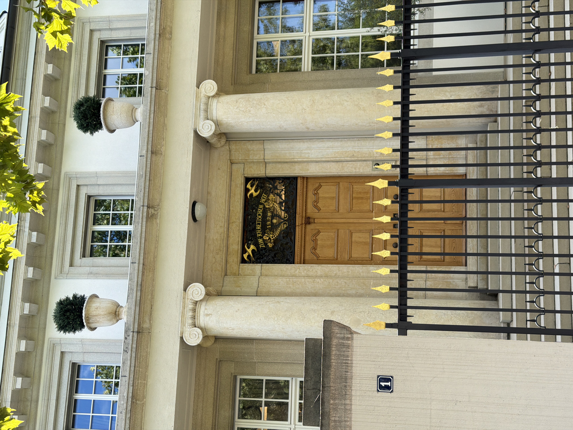 The image depicts the facade of the Rätisches Museum in Basel, Switzerland, a building characterized by its neoclassical architecture. The building features a prominent entrance with double wooden doors, framed by two large Ionic columns. Above the entrance, a decorative wrought-iron grille is mounted, displaying the inscription "ZUM RÄTISCHEN HOF 1325". On the upper level of the building, several large windows with white frames are visible, interspersed with decorative urns containing small green trees. The foreground is dominated by a black wrought-iron fence with gold decorative elements at the top. A small plaque with the number "1" is affixed to a light-colored wall to the left of the fence. The building is bathed in sunlight, and lush green leaves from trees are visible in the upper part of the frame, suggesting a daytime scene in either spring or summer.