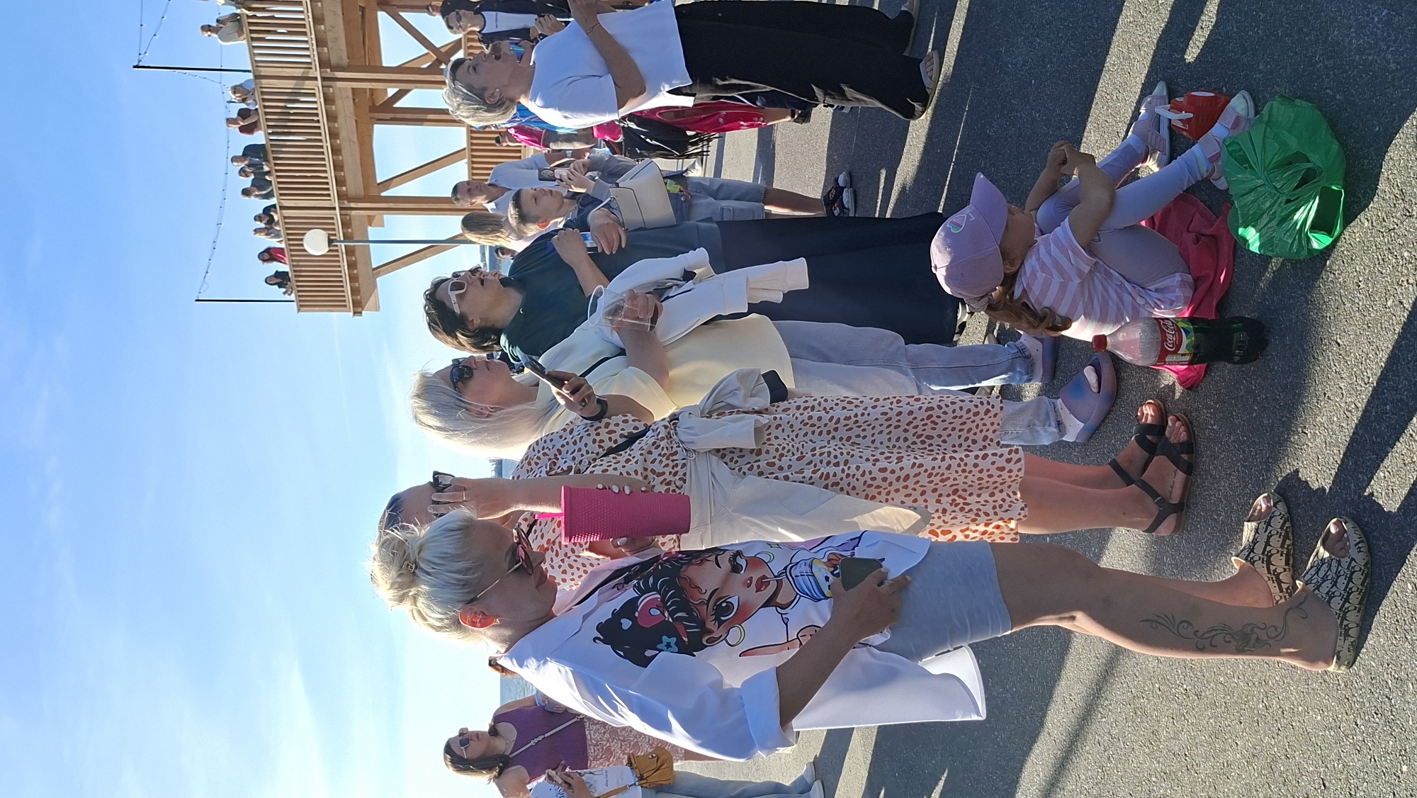 In Tallinn, Estonia, a group of people stand on a paved surface near a body of water under a clear blue sky. In the foreground, a woman with blonde hair and sunglasses wears a white oversized shirt with a cartoon graphic and grey shorts, holding a pink cup. Next to her, another woman in a patterned dress and black sandals stands holding a phone. To her right, a woman in a dark green hooded top and black pants is visible. Further to the right, a young boy holds a phone. In the background, a wooden structure with multiple levels and railings is present, with people standing on the upper levels, overlooking the scene. A little girl in a pink striped shirt and a purple hat sits on the ground to the right, next to a bottle of Coca-Cola and a green plastic bag. The scene appears to be an outdoor gathering or event.