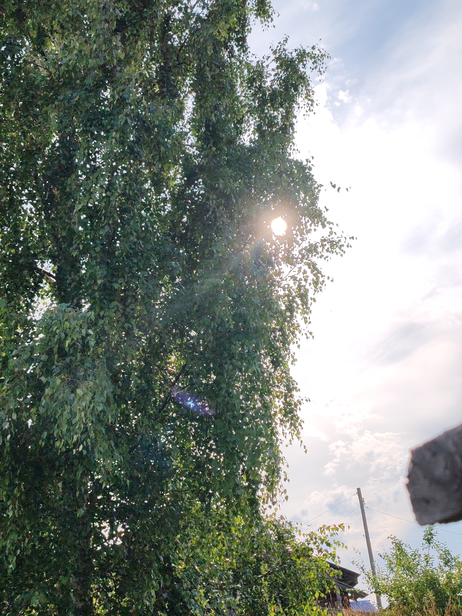 A close-up, low-angle shot captures a dense canopy of green leaves from a birch tree, with the sun piercing through the foliage in the upper center. Lens flare, appearing as a bright white circle and a blue and purple glint, is visible. The sky, with scattered white clouds, occupies the upper right quadrant. In the background, a utility pole with wires and a roofline of a building are partially visible. The lower right corner shows a rough, gray concrete object, possibly part of a structure or debris. The scene is likely outdoors in a rural or suburban setting, possibly in or near Чухлэм, Russia, suggested by the overall aesthetic and vegetation.