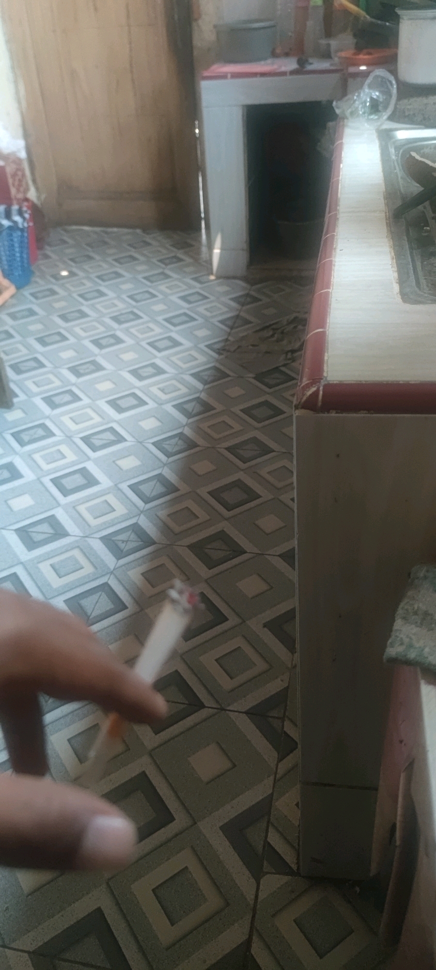 The image shows a close-up of a person's hand holding a lit cigarette over a patterned tiled floor. The floor has a repeating diamond design in shades of grey and white. In the background, there is a wooden door on the left and a counter with a sink on the right. The scene appears to be indoors, possibly a kitchen or utility area. There is no visible indication of the location being in Gambarsari, Indonesia.