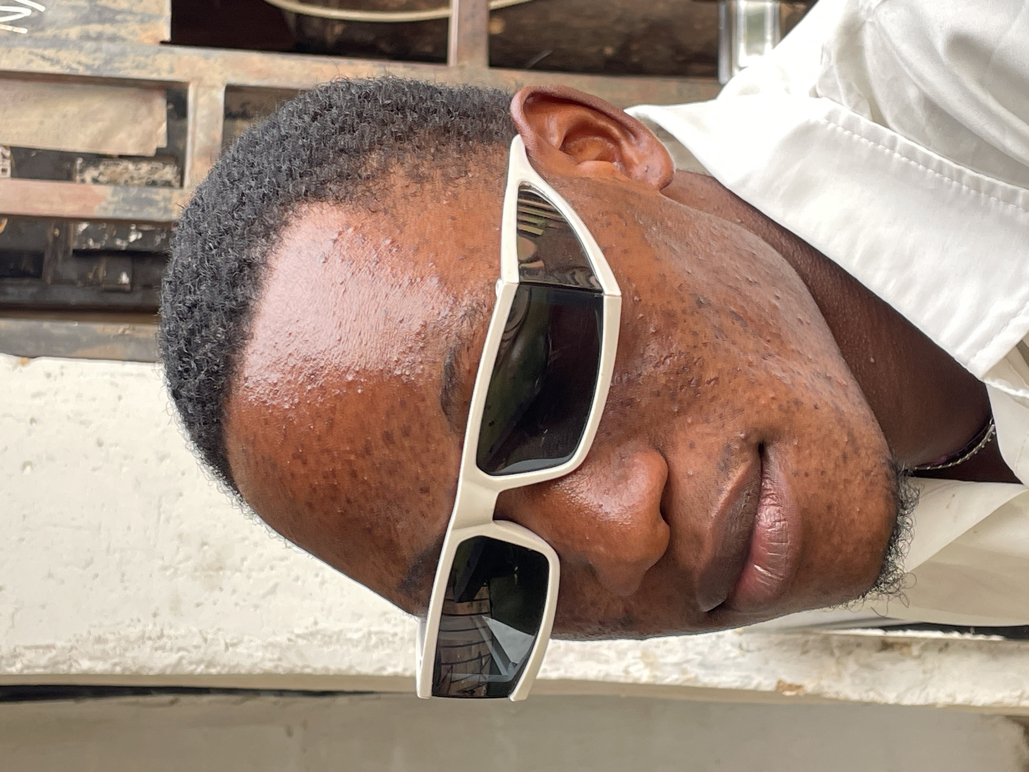 A dark-skinned man with short, curly black hair and white-framed sunglasses is pictured outdoors, likely during the day, as evidenced by the natural lighting. The man is wearing a white shirt, and a silver necklace is visible. The setting appears to be a relatively nondescript outdoor location, with a metal structure with horizontal bars behind the man's head. There is no explicit evidence to indicate the location as Jos, Nigeria.