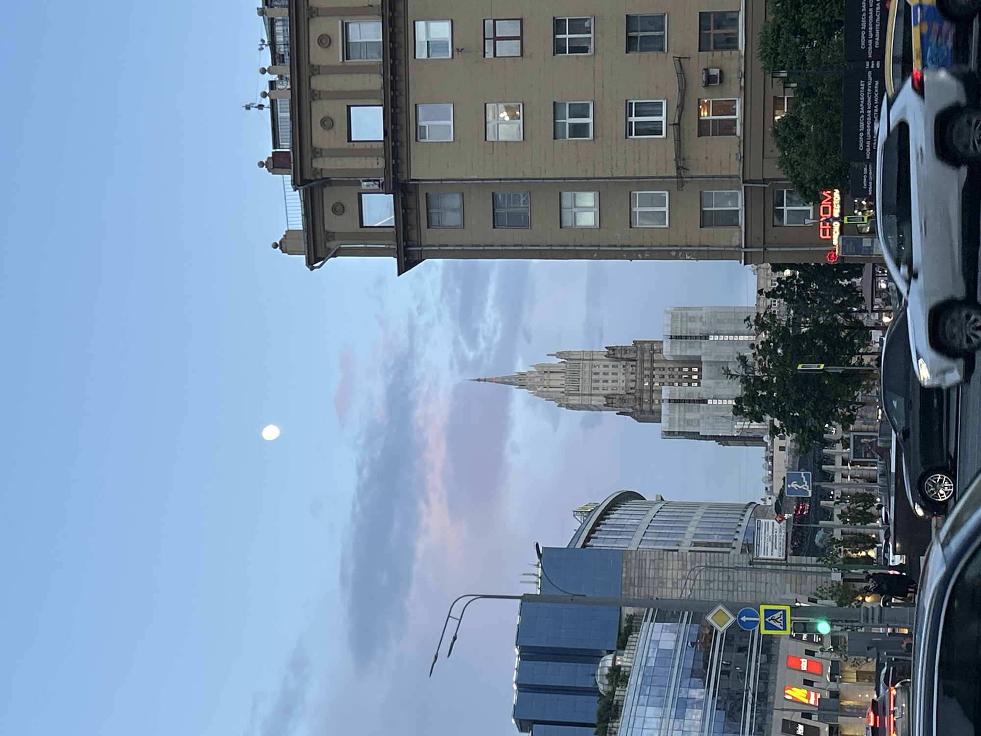 The image shows a cityscape with a mix of old and modern buildings under a sky with a moon. The buildings include a light brown structure with multiple windows, likely an older residential or office building. In the distance is a taller, more ornate building with a spire, possibly a government or historical landmark. In the foreground are modern glass and steel structures, indicative of a business or commercial district. Street signs and traffic lights are visible, and there are cars parked along the side of the road. There are also signs in Russian, which say, "FROM" "PLACE RESTAURANT" and "SOON THERE WILL START WORKING" "NEW DIGITAL STRUCTURE IN" "GOVERNMENT OF MOSCOW". The image appears to be taken from a ground-level perspective, likely from a street. The location appears to be Moscow.