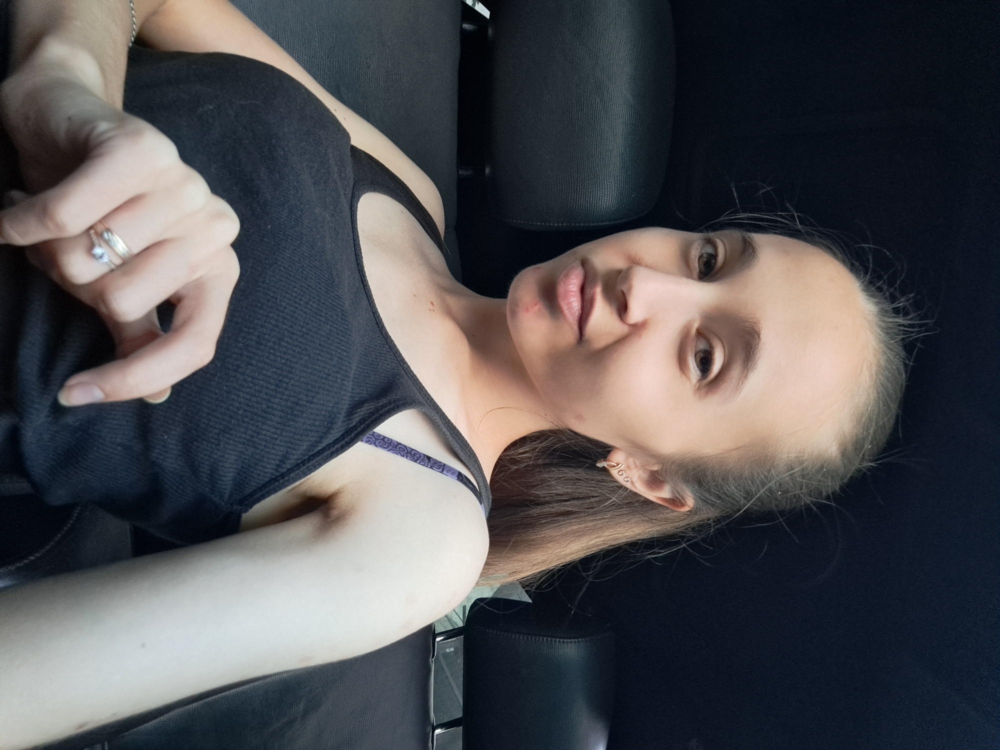 A young woman with light brown hair pulled back in a ponytail is sitting in the backseat of a car. She is wearing a black tank top and a patterned bra strap is visible. She has earrings in her left ear and rings on her right hand. She is looking at the camera and her right hand is resting on her chest. The background is dark and appears to be the interior of a car, with black leather seats visible. The image does not provide enough information to determine the location within Vypasne, Ukraine.