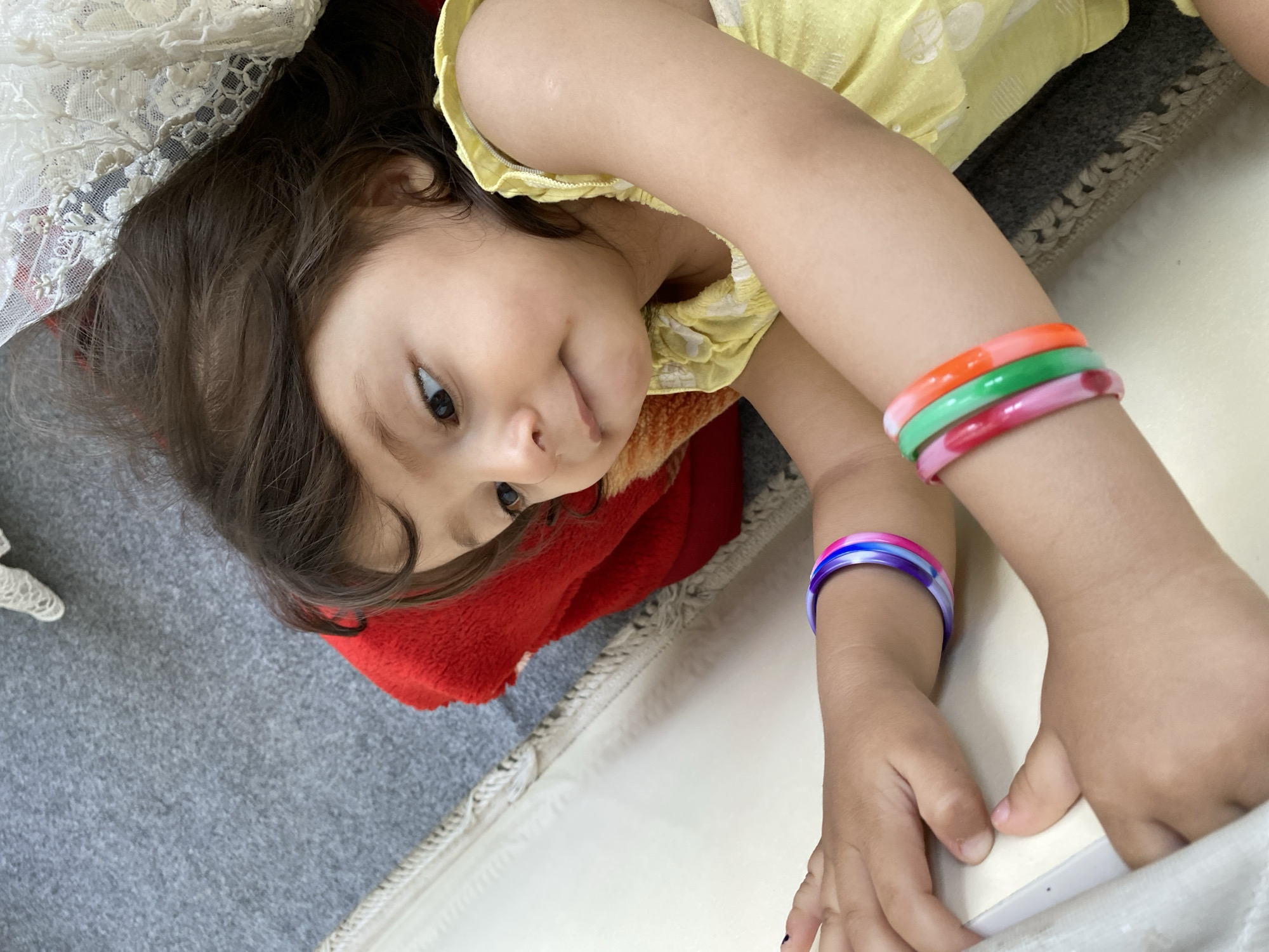 A young child, likely a girl, is captured in a close-up, slightly tilted shot, lying down indoors in Kandahar, Afghanistan. She has dark brown hair and observant brown eyes, looking towards the viewer with a gentle, slightly smiling expression that conveys a sense of quiet contentment or curiosity.
She is wearing a light yellow top, possibly patterned. Her arms are raised, displaying multiple vibrant bangles. On her right wrist (from the viewer's perspective), there are three bangles: one orange, one green, and one pink/red with white swirls. Her left wrist is adorned with a single multi-colored bangle featuring shades of purple, blue, pink, and white. The fingernails on her left hand have a dark polish.
The child is resting on a light-colored surface, possibly a blanket or rug, with a darker grey carpet visible in the background. A white, lacy fabric, which could be part of a curtain or bedding, frames the upper left corner of the image. A red, fluffy blanket or garment is tucked underneath her head and shoulder area, adding a splash of contrasting color. The lighting suggests an indoor setting, possibly during the daytime. The image focuses intimately on the child, highlighting her innocent expression and colorful accessories.