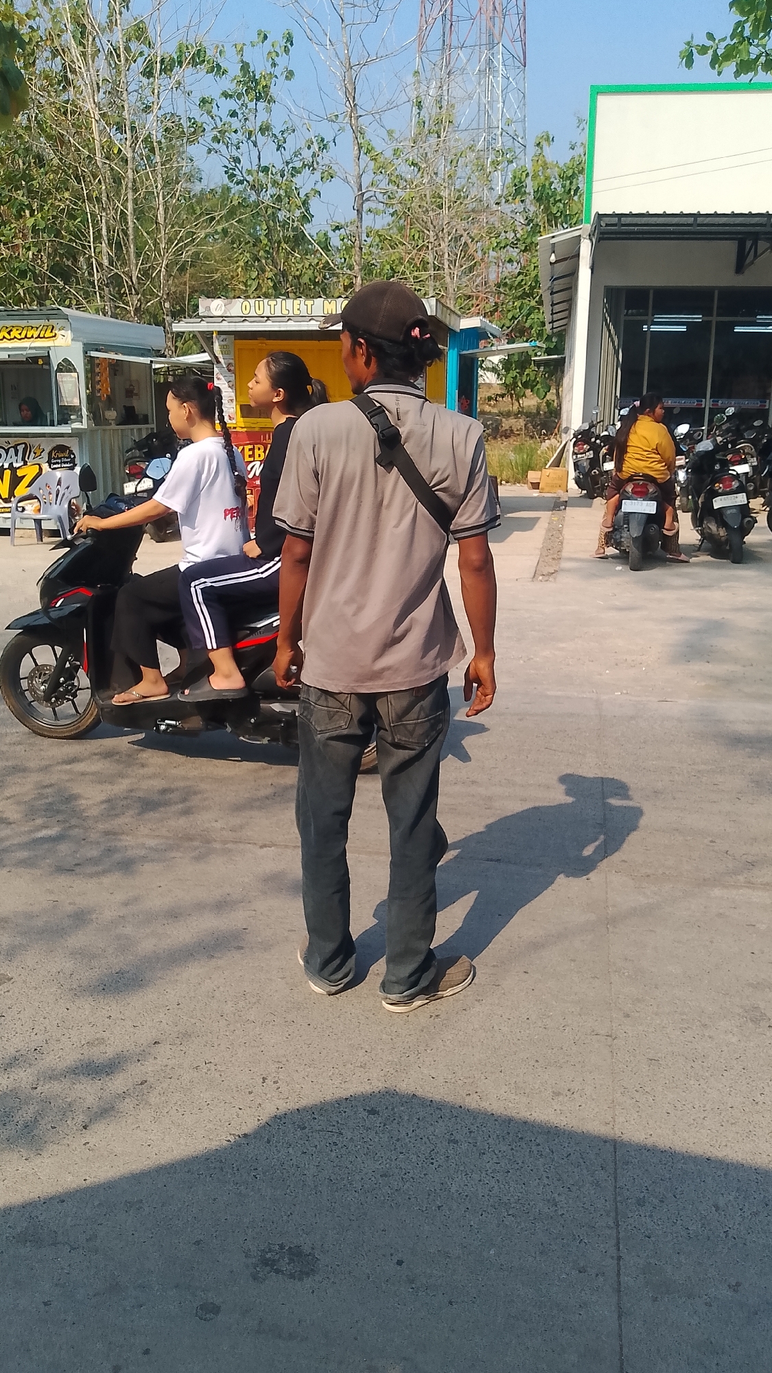 The image depicts a street scene in Karanglangu, Indonesia, under a clear sky. In the foreground, a man wearing a grey t-shirt and dark jeans stands facing away from the camera, with a black crossbody bag slung over his shoulder. His shadow stretches across the asphalt. Behind him, two young women are seated on a black motorcycle. The woman in front is wearing a white t-shirt and black pants, while the woman behind her is wearing a black outfit. To the left of the motorcycle, there are food stalls with signs, one of which reads "KRIWIL" and another "OUTLET M". In the background, several other motorcycles are parked, and a building with a green trim is visible. The environment appears to be an outdoor area with trees and a telecommunications tower in the distance.