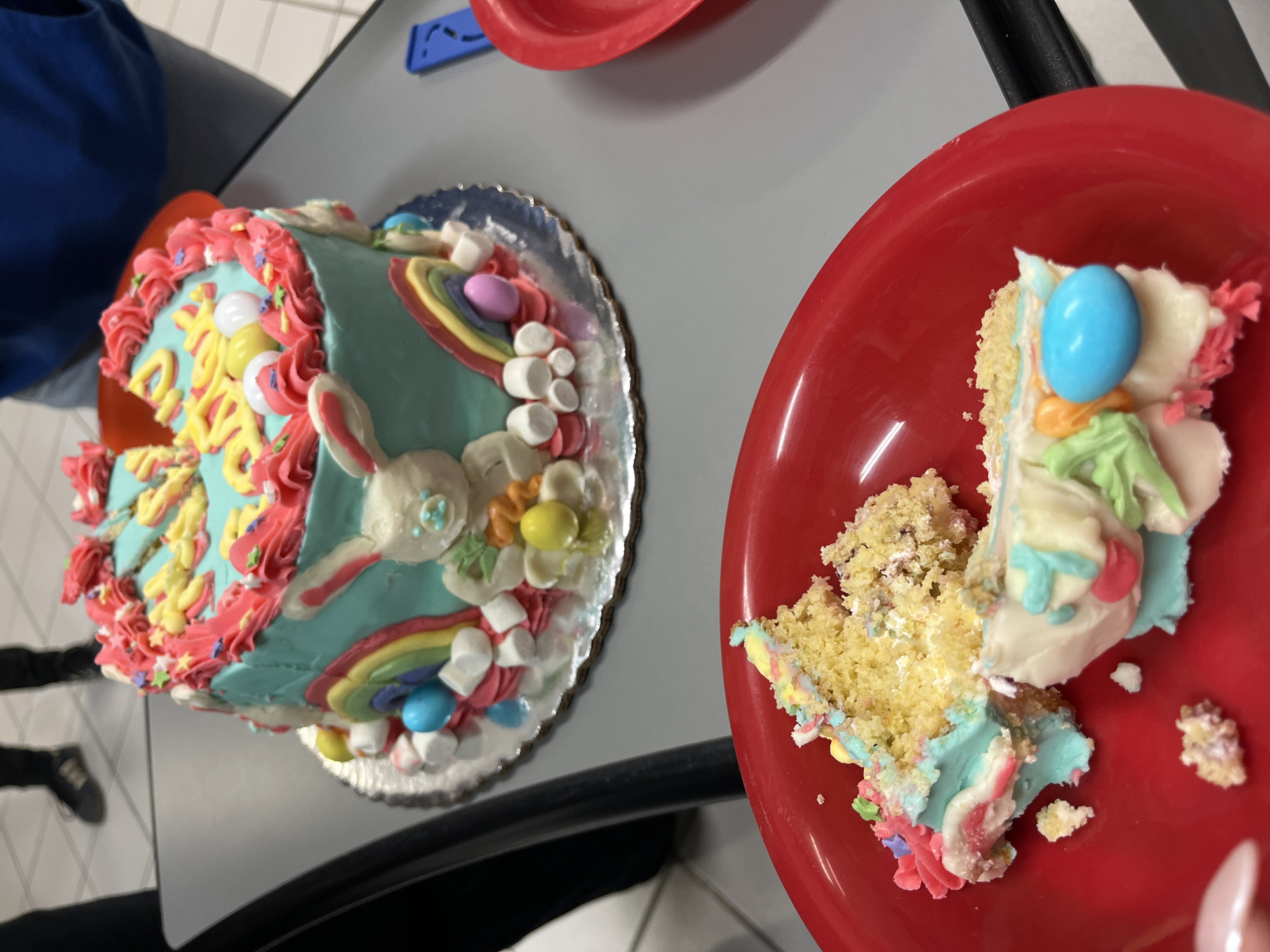 The image depicts an indoor scene focused on two Easter-themed cakes. In the midground, a whole, round, two-tier cake rests on a silver scalloped cake board placed on a smooth, gray table. The cake is predominantly light blue and features "Happy Easter" spelled in yellow letters on top, surrounded by pink frosting rosettes and multicolored sprinkles, including small stars. The sides of the cake are decorated with white frosting bunnies, rainbow arches, white marshmallows, and various pastel-colored candy eggs (yellow, blue, pink, white). To the right and slightly behind this cake, a red plastic bowl is partially visible, along with a blue rectangular object on the gray table surface.
In the foreground, a bright red plastic bowl holds several partially consumed pieces of a cake slice. This cake exhibits a yellow sponge base with light blue, pink, white, and green frosting. A prominent bright blue candy egg is visible among the frosting on one piece, alongside other smaller pastel candies. The lower left of the image shows the edge of a person's hand, specifically a thumb with a light-colored nail, near the red bowl.
The background reveals a tiled floor composed of white and gray squares. The lower legs and feet of at least two individuals are visible: one person wears black pants and black shoes on the left, and another person wears blue pants and a blue top on the right. The lighting suggests an artificial indoor environment. The specific location within the city of Puebla, Mexico, is not discernible from the visual information provided.