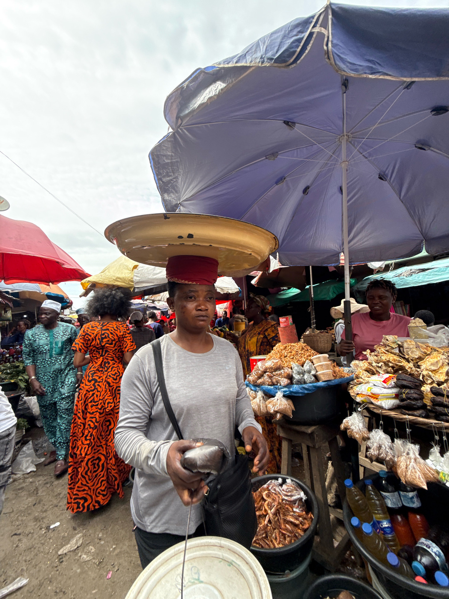 This is a medium shot of a bustling marketplace in Lagos, Nigeria, under an overcast sky. The foreground is dominated by a woman wearing a grey long-sleeved shirt and dark pants, with a black bag slung across her body. She is balancing a large, round, golden tray on her head, secured by a red fabric wrap. Her hands are clasped, and she appears to be holding a metallic ladle.
Behind her, the market is alive with activity. To her left, a woman in a vibrant orange dress with a black wavy pattern is walking away from the camera. Further to the left, a man in a green and teal patterned traditional outfit and a white cap is also walking. Various stalls are visible, adorned with colorful umbrellas, including a large blue one partially obscuring the sky, and a red one to the far left.
The stalls display a variety of goods, including what appears to be dried fish and shrimp, packaged food items, and bottles of liquid, possibly drinks. The ground is dusty and uneven, with some scattered debris. The overall impression is of a lively and commercial environment, typical of a Lagos market.