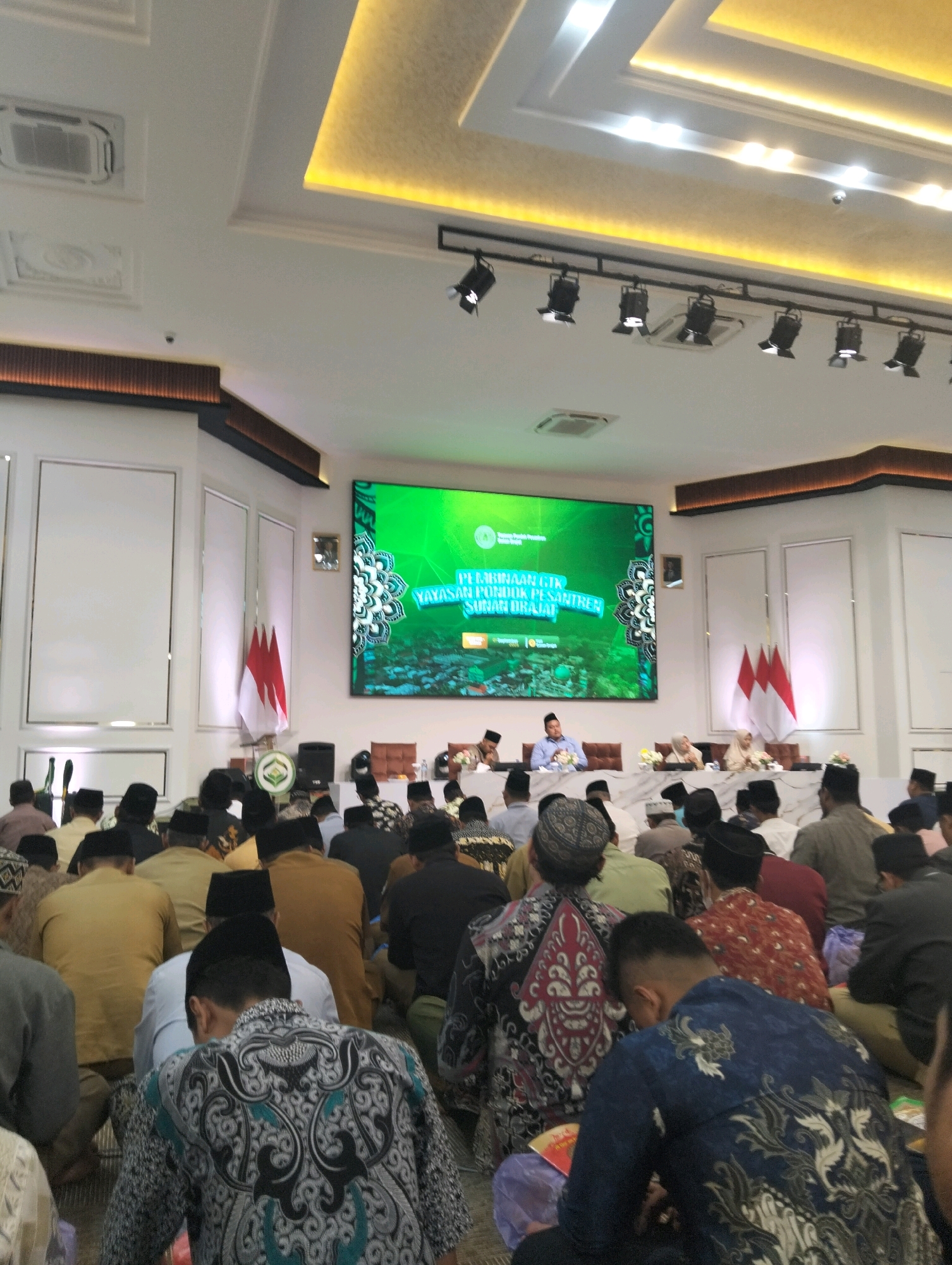 In a well-lit indoor venue, a large screen displays a green background with white text and intricate mandala-like patterns, featuring the words "PELATIHAN GTK" and "YAYASAN PONDOK PESANTREN SURAH AL-AQSA." Several people are seated at tables on a stage in front of the screen, appearing to be speakers or honored guests. Three Indonesian flags, red and white, are displayed behind them. Below the stage, a large audience of men, mostly wearing traditional Islamic attire such as peci (caps) and sarongs or batik shirts, are seated on the floor, facing the stage. The environment appears to be a conference or religious gathering. The presence of the Indonesian flag and attire suggests a location within Indonesia, possibly at a pesantren (Islamic boarding school) or a related institution. The image does not provide enough specific visual cues to pinpoint the exact city of Dagan, Indonesia.