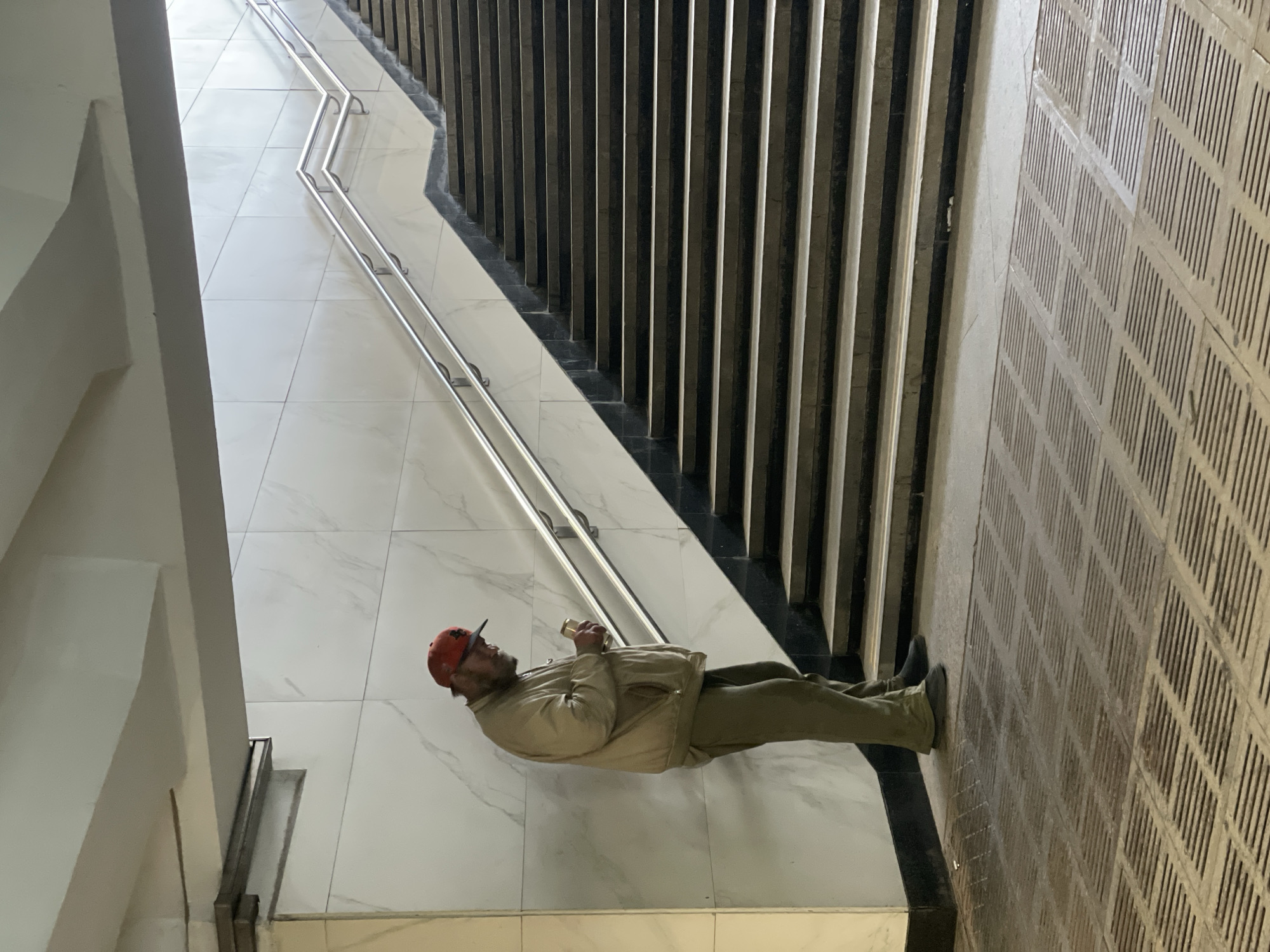 A middle-aged man stands indoors within a modern architectural space. He wears a red baseball cap, tan jacket, and olive green pants. He holds a small cylindrical object, possibly a can or cup, in his right hand. He is positioned on a light-colored tile floor with a marble-like pattern. A stainless steel handrail runs alongside a set of stairs with a black trim. Adjacent to the stairs are vertical, dark-colored linear elements, possibly part of a decorative screen or wall. A patterned wall is on the right side of the frame. Based on visual analysis and the provided location, this is likely a public area in Khimki, Russia, such as a subway station or shopping mall.