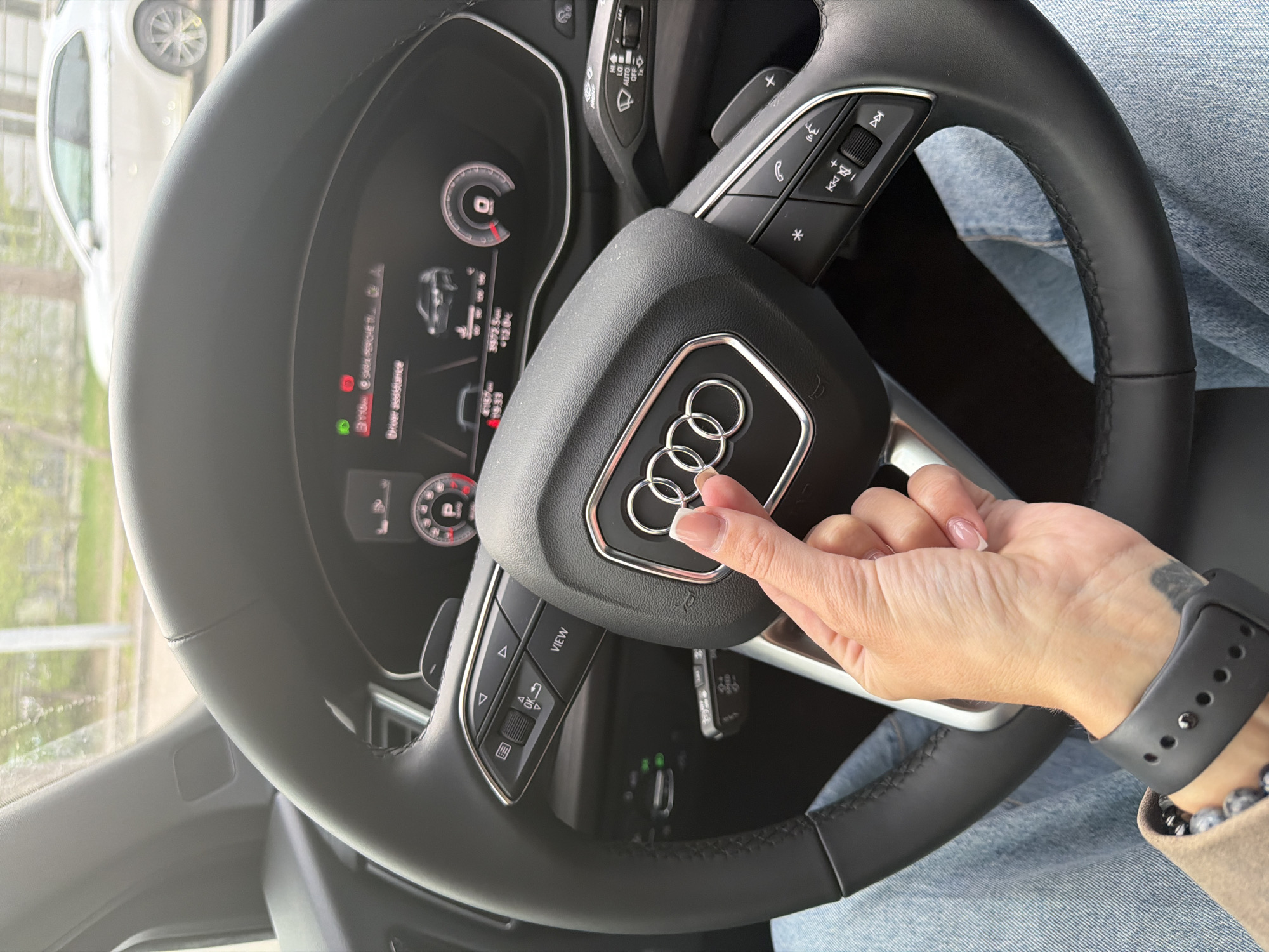 A close-up shot shows the steering wheel of an Audi car, with a woman's hand touching the Audi logo in the center. The hand wears a black smartwatch and has a light-colored nail polish with a white tip. The car's dashboard is partially visible, displaying various driving information. The steering wheel has multiple buttons and controls. A blurred image of a white car can be seen through the windshield. The interior of the car is visible. The location appears to be Unknown, Unknown.