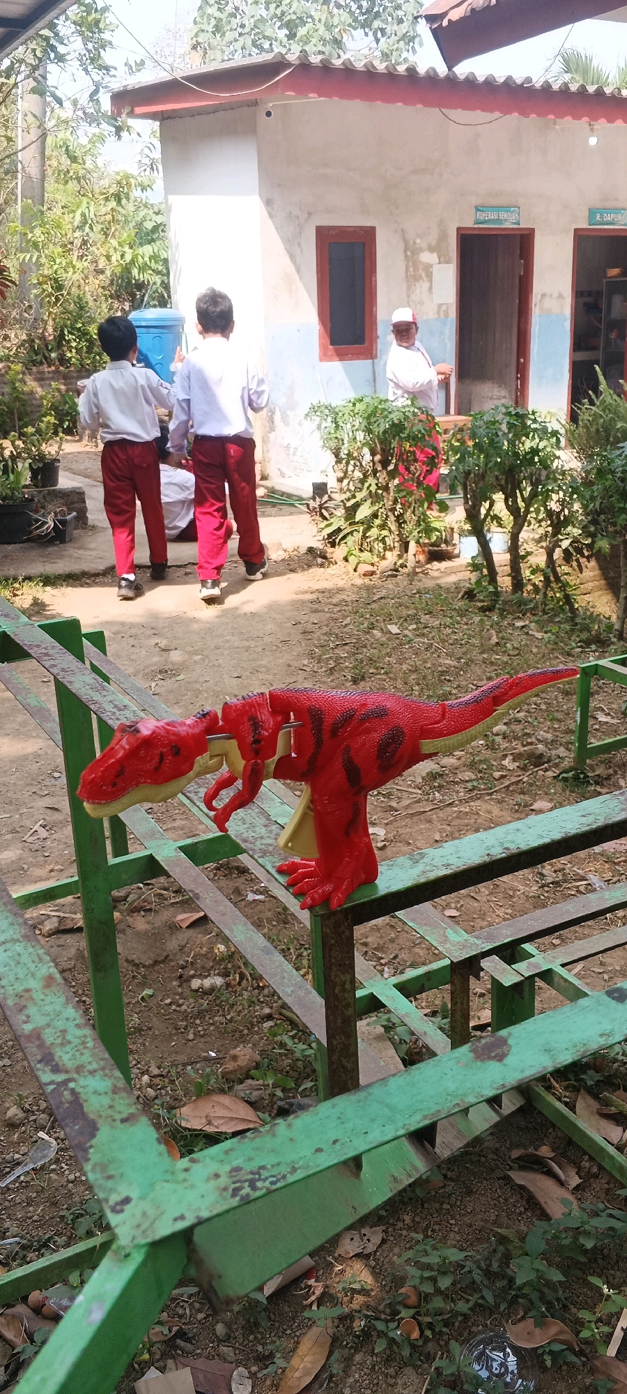 In an outdoor setting in Penanggungan, Indonesia, a red plastic toy dinosaur is perched on a rusted green metal railing. The dinosaur is in a dynamic pose, with its mouth open. In the background, two young boys in white shirts and red pants are walking away from the camera. A third child, also in similar school attire, is partially visible sitting on the ground. Behind them is a light-colored building with a red-tiled roof, featuring two doorways with signs indicating "KOPERASI SEKOLAH" (School Cooperative) and "R. DAPUR" (Kitchen). A white-clad adult wearing a cap stands near one of the doorways. The ground is a mixture of dirt and dry leaves, with some greenery and potted plants scattered around. The scene suggests a school or community area on a sunny day.