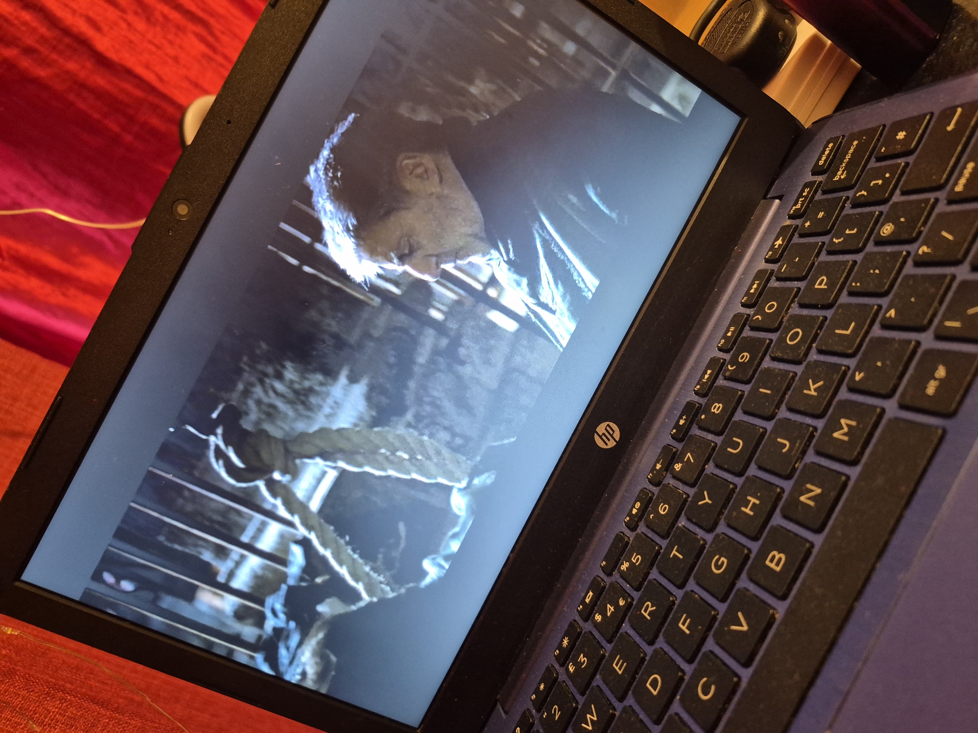 A laptop screen displays a scene from a movie. In the foreground, a thick rope hangs from the top of the frame, suggesting a scene of potential distress or historical context. A man, seen in profile, looks downwards with a weary expression. He has short, graying hair and appears to be wearing dark clothing. The background of the screen is dark and appears to be a dimly lit interior with wooden structures, possibly bars or scaffolding. The laptop itself has a dark keyboard with illuminated keys, showing the familiar QWERTY layout, and the HP logo is visible on the right side of the keyboard area. The laptop is placed on a surface covered with a red textured material, possibly a blanket or tablecloth. A thin yellow wire is visible extending from the top of the frame. The image is taken from a slightly elevated angle, looking down at the laptop. The context of Sandwell, United Kingdom, is not present in the visual information.