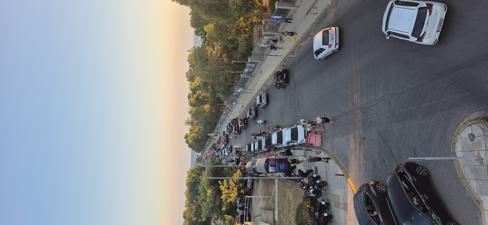 The image depicts a street scene in Paleo Faliro, Greece, during what appears to be late afternoon or early evening, given the warm lighting and clear sky. A considerable amount of traffic, composed primarily of cars and some motorcycles, is visible on a multi-lane road that curves gently. The road is flanked by a sidewalk on the left and a tree-lined area and a fence on the right.
On the left sidewalk, several people are standing or walking. There are also parked motorcycles and a white bus stop shelter. Some cars are parked along the left side of the road.
The road itself has traffic moving in both directions, with some congestion. Several pedestrians are visible on or near the sidewalk.
The right side of the road features a pedestrian walkway with a fence separating it from a densely wooded area. More people are seen walking on this side. In the distance, beyond the trees, the sea is visible, with a few boats or ships on the horizon.
The overall impression is that of a busy urban coastal area during a period of high activity.