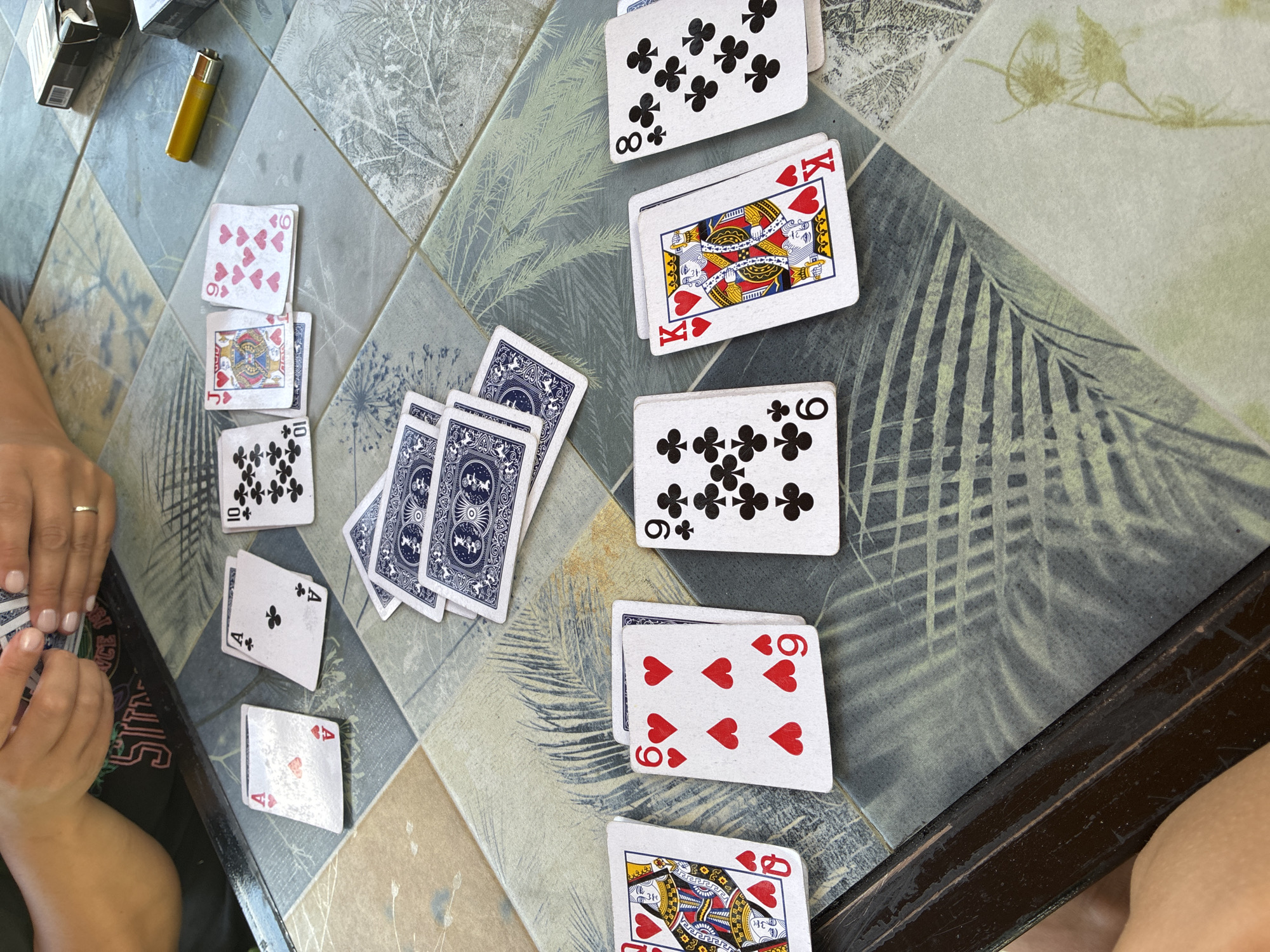 In Surami, Georgia, a group of individuals is engaged in a card game around a tiled table. The table's surface features a pattern of beige, blue, and grey tiles, some adorned with abstract botanical designs in grey and green. Scattered across the table are various playing cards, predominantly from a standard deck with red hearts and black clubs. Visible cards include an Ace of Hearts, Ace of Clubs, a 10 of Clubs, a Jack of Hearts, a 9 of Hearts, a 6 of Hearts, a King of Hearts, and an 8 of Clubs. Two decks of cards, one with a blue and white patterned back and another with a predominantly blue design, are also present, with one person's hands seen shuffling or holding cards. A yellow lighter and two cigarette packs, one partially open, are placed near the cards. The setting appears to be outdoors, possibly a patio or balcony, given the ambient light and the texture of the table.