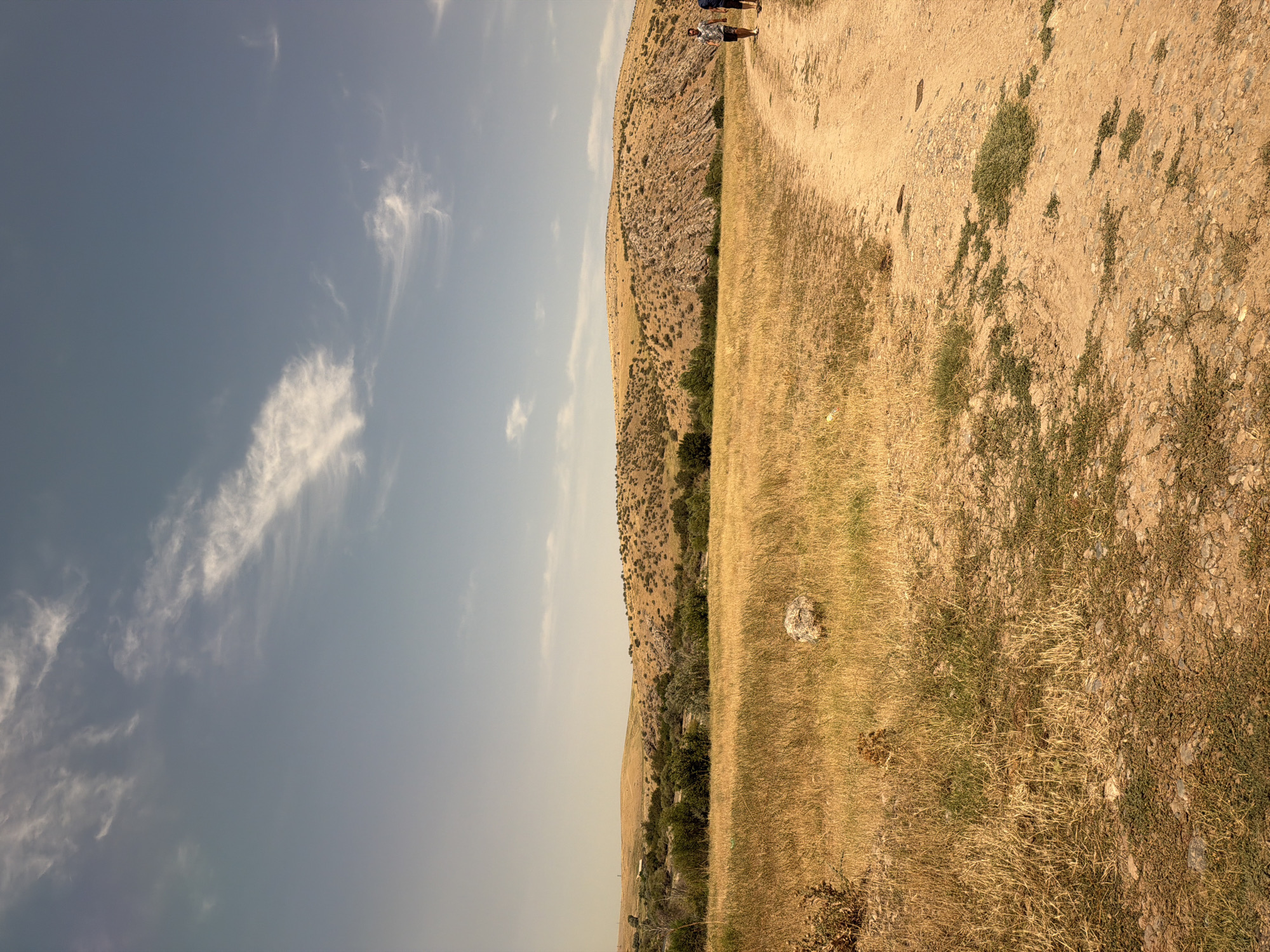 Two individuals are walking on a dirt path in a dry, grassy landscape with hills in the background. The sky is clear with a few wispy clouds. The scene is likely located in the vicinity of Boraldai, Kazakhstan, known for its steppe environment.