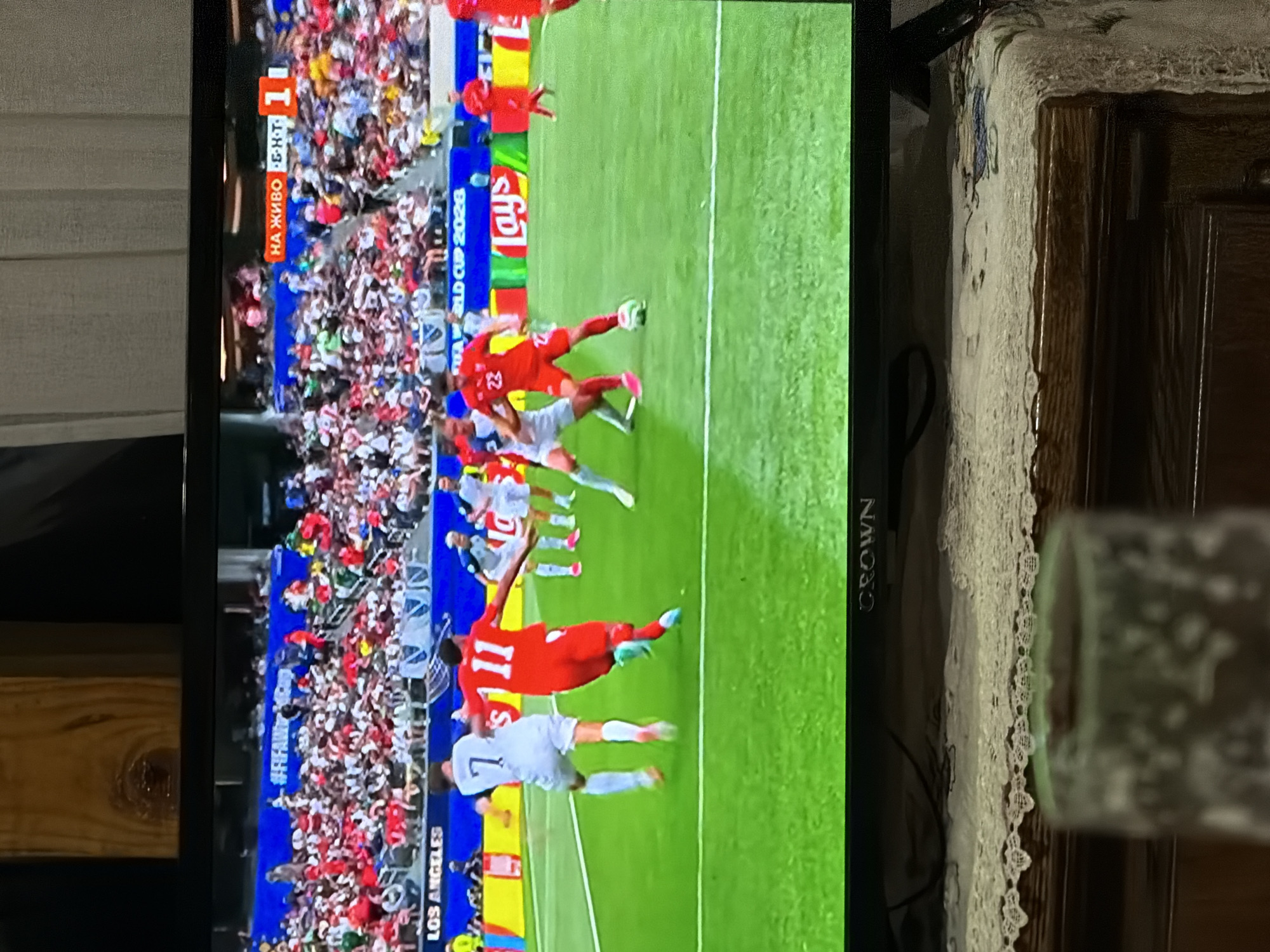 In the city of Lobosh, Bulgaria, a television screen displays a live soccer match. The game is being broadcast on what appears to be a large flat-screen television. The screen shows a green soccer field with several players in red and white uniforms actively engaged in the game. In the background, a large crowd of spectators is visible in the stands, dressed in a mix of red, white, and green clothing, suggesting a nationalistic fervor or team support.
The top of the screen displays broadcasting information, including "НА ЖИВО" (LIVE) and a score indicator of "1" within a red square. Below the field, a banner reads "FIFA WORLD CUP 2026" and the "LOS ANGELES" logo, suggesting the broadcast is for a World Cup qualifying match or a related event being hosted in Los Angeles. The television is placed on a wooden dresser covered with a white lace cloth, and a partially filled glass with frothy liquid sits in the foreground, obscuring the bottom portion of the image. The overall scene captures a moment of sports viewing at home, with the backdrop of a live international soccer event.