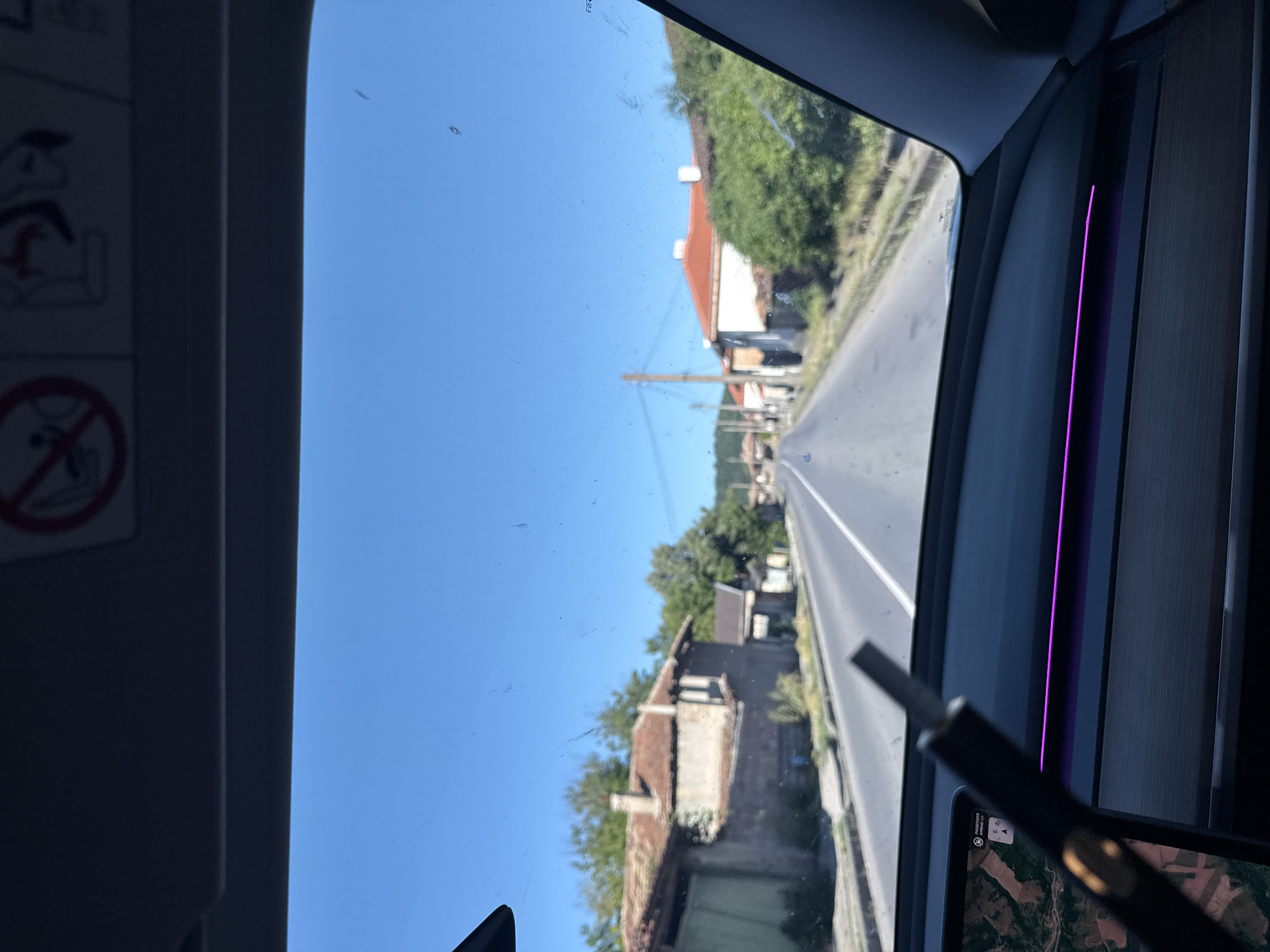 The image shows a view from inside a vehicle traveling on a paved road in the village of Veselinovo, Bulgaria. The road is flanked by traditional houses with red-tiled roofs and weathered facades. Green trees and vegetation line the sides of the road and surround the buildings, indicating a rural or semi-rural setting. The sky is clear and blue, suggesting a sunny day. The interior of the vehicle is visible in the foreground, with a dashboard and a digital display showing a map. A dark object, possibly a pen or stylus, is held in front of the screen. The overall scene depicts a journey through a Bulgarian village on a bright, clear day.
