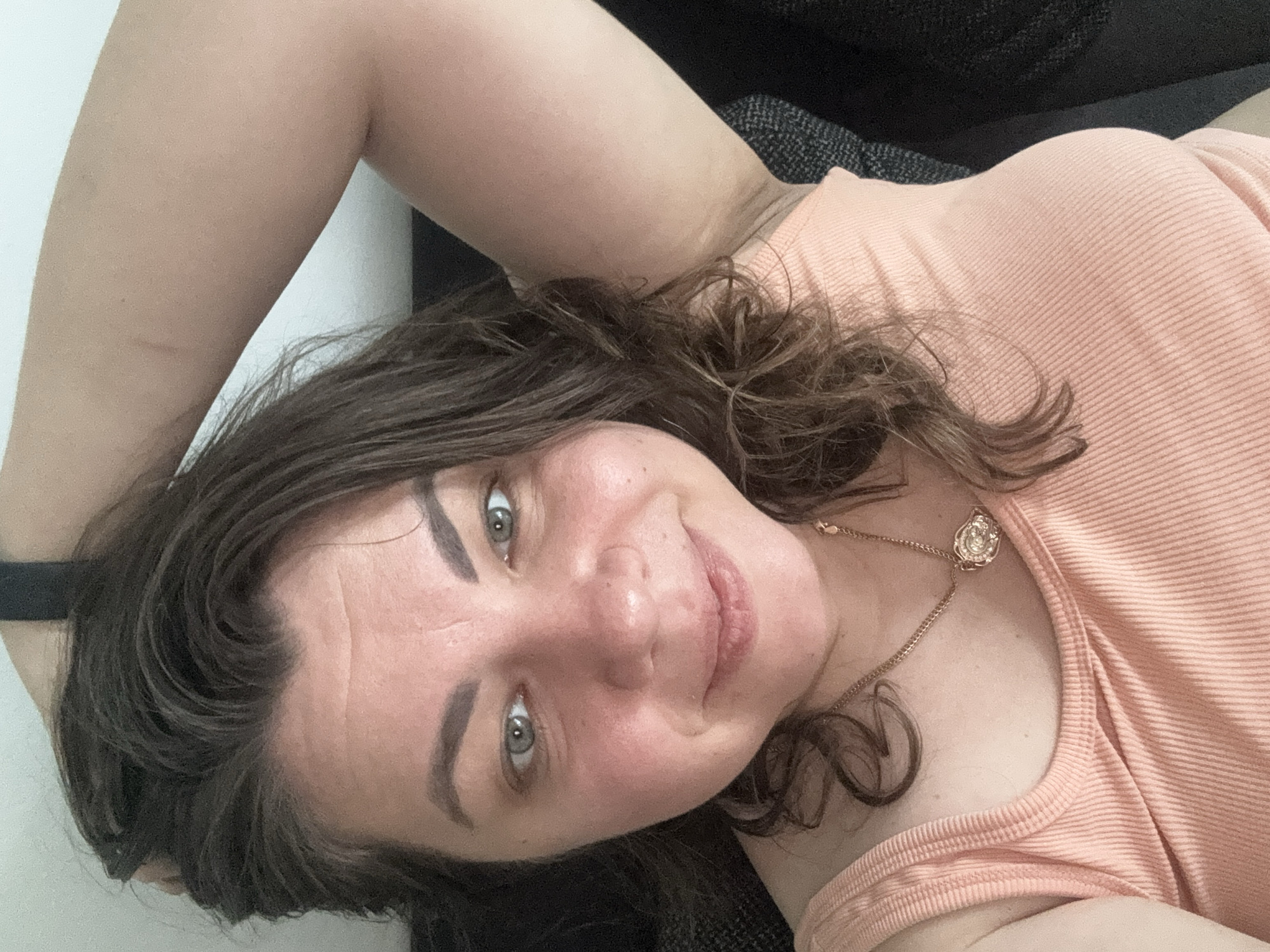 A woman with brown hair and green eyes, wearing a peach-colored tank top and a gold necklace, is captured in a close-up, over-the-shoulder selfie. Her right arm is raised, resting her hand on the back of her head. She is indoors, likely at her residence in Höhenkirchen-Siegertsbrunn, Germany, as indicated by the background which shows a lightly colored wall and a dark-colored couch. The lighting appears to be natural, coming from a window or overhead source, casting soft shadows on her face. The image is taken from a slightly high angle, looking down at the woman.