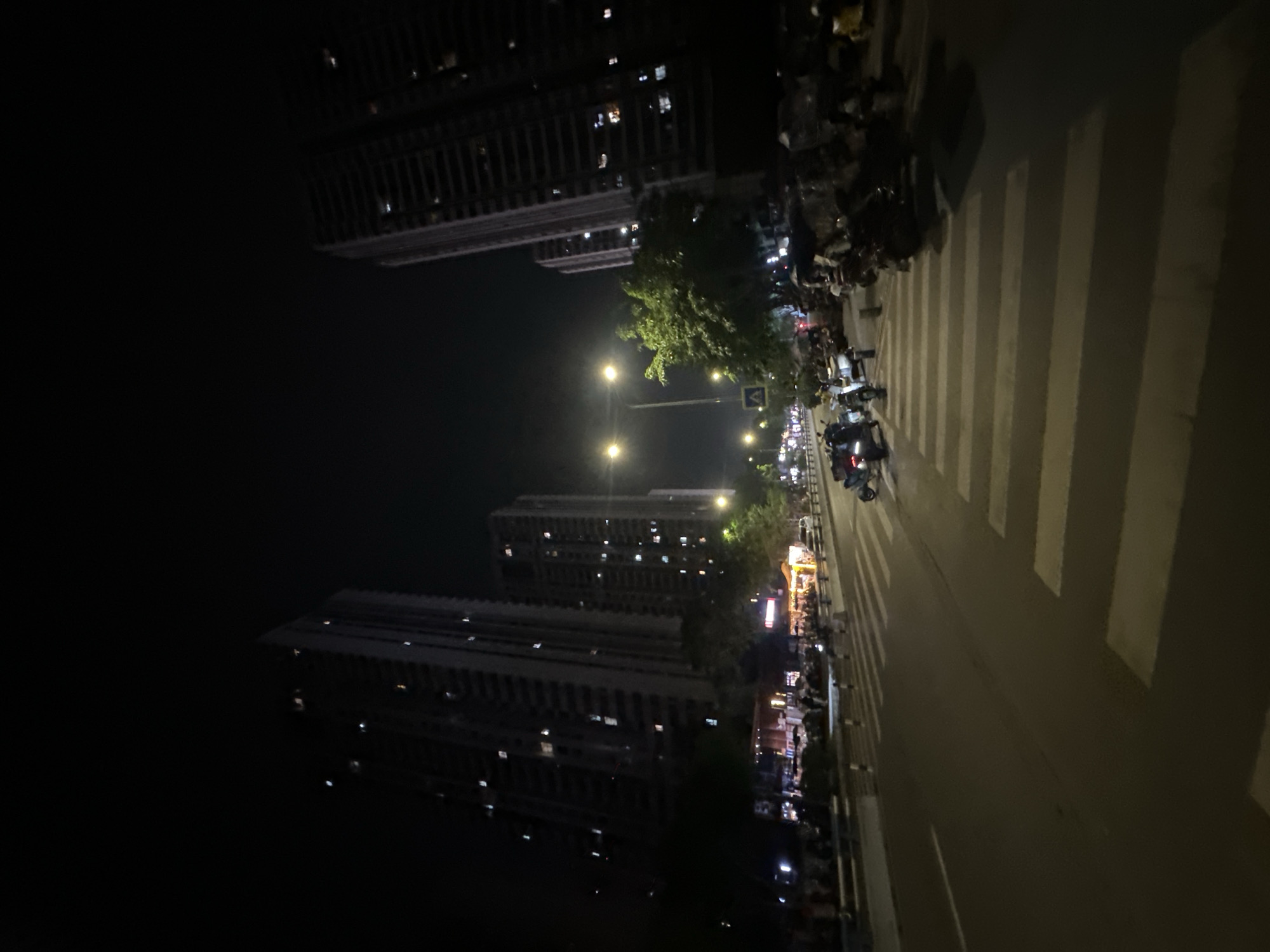 The image depicts a nighttime street scene in the Chongqing High-tech Zone, China. Tall, illuminated apartment buildings dominate the background, with numerous windows lit. In the foreground, a wide, asphalt road features a prominent pedestrian crossing marked with white stripes. Several scooters and motorbikes are parked along the side of the road and on the sidewalk, with some individuals visible near them. Shops and food stalls with illuminated signs and awnings line the street on the left side, indicating a commercial area. Trees are interspersed along the sidewalk. Streetlights cast a warm glow on the road and surrounding environment. The overall atmosphere is that of a busy urban area at night.