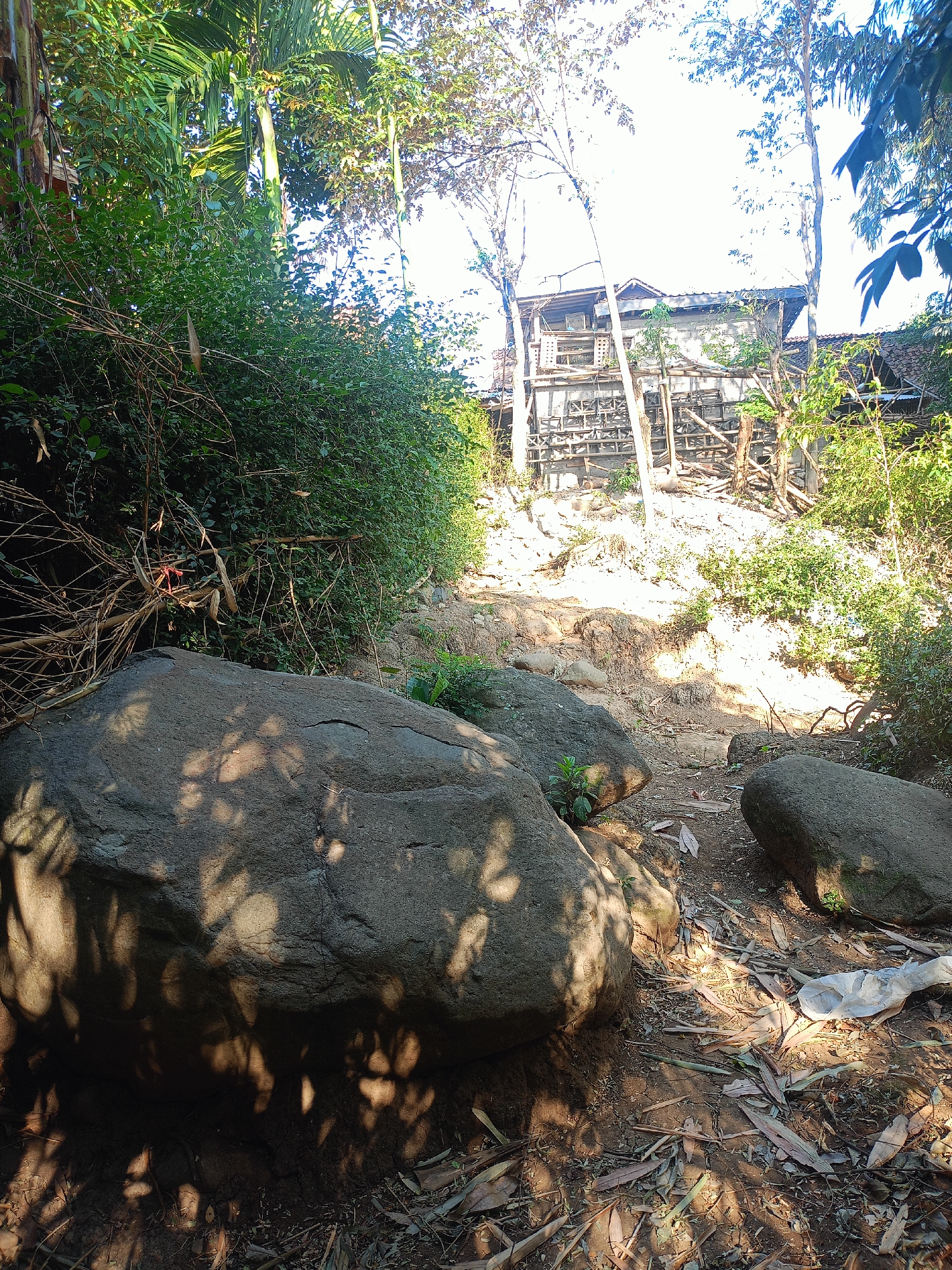 This image depicts a rural scene in Jelbuk, Indonesia, with a focus on a natural pathway leading towards a partially constructed or dilapidated building. In the foreground, large rocks are scattered along a dirt path covered with fallen leaves and debris. Lush green foliage and trees line the left side of the path, casting dappled shadows. In the midground, the path slopes upward towards a structure characterized by exposed wooden scaffolding and unfinished walls. The building appears to be situated on a higher elevation, surrounded by vegetation. The sky is visible in the background, suggesting an outdoor setting under daylight. There are no people or animals present in the image.