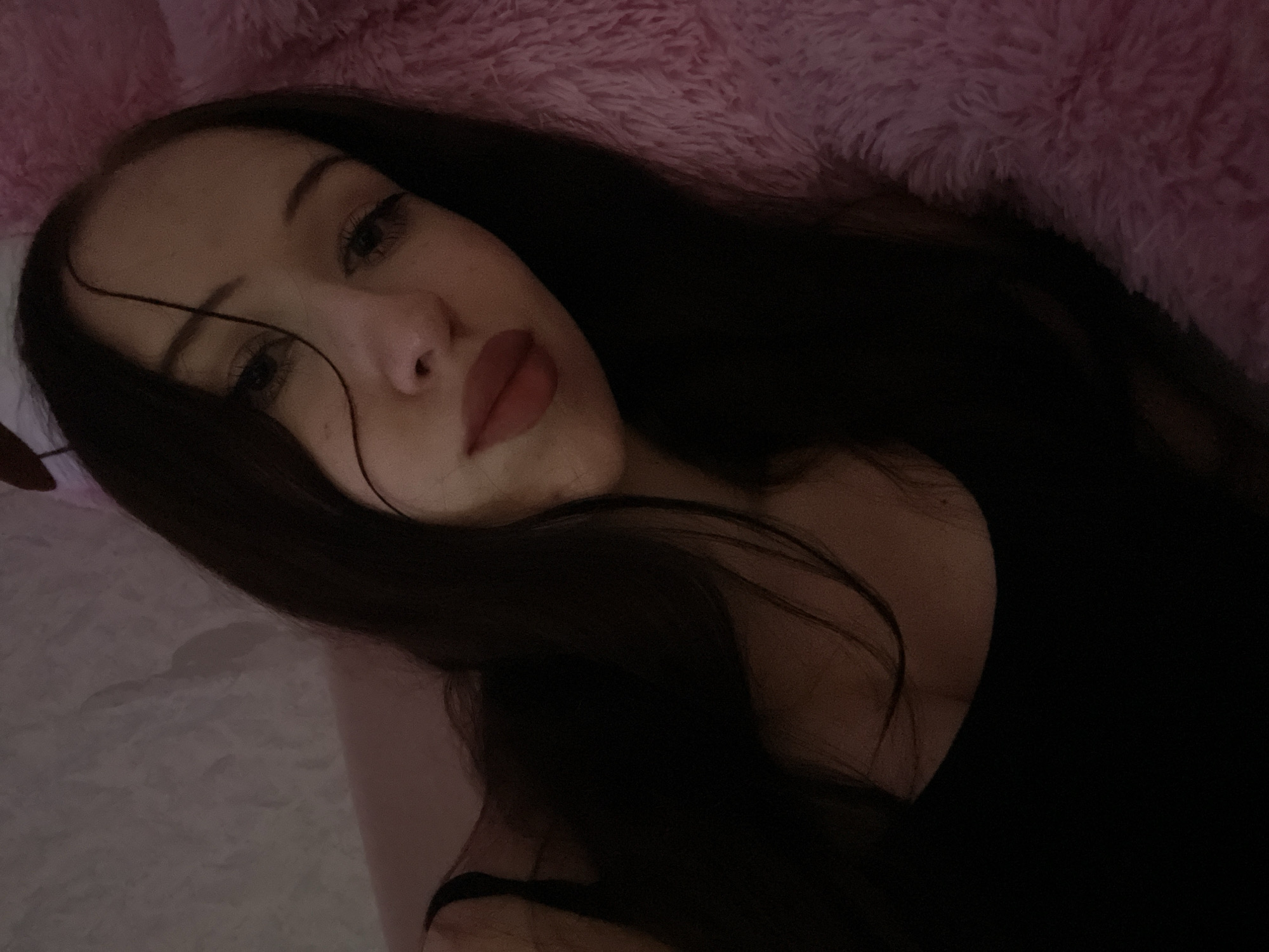 A young woman with dark hair and full lips lies in bed, illuminated by soft lighting. She wears a black tank top and her hair is spread across her shoulders and the pink, fluffy blanket she rests on. The background is a textured, light-colored wall. The scene suggests a private, intimate moment.