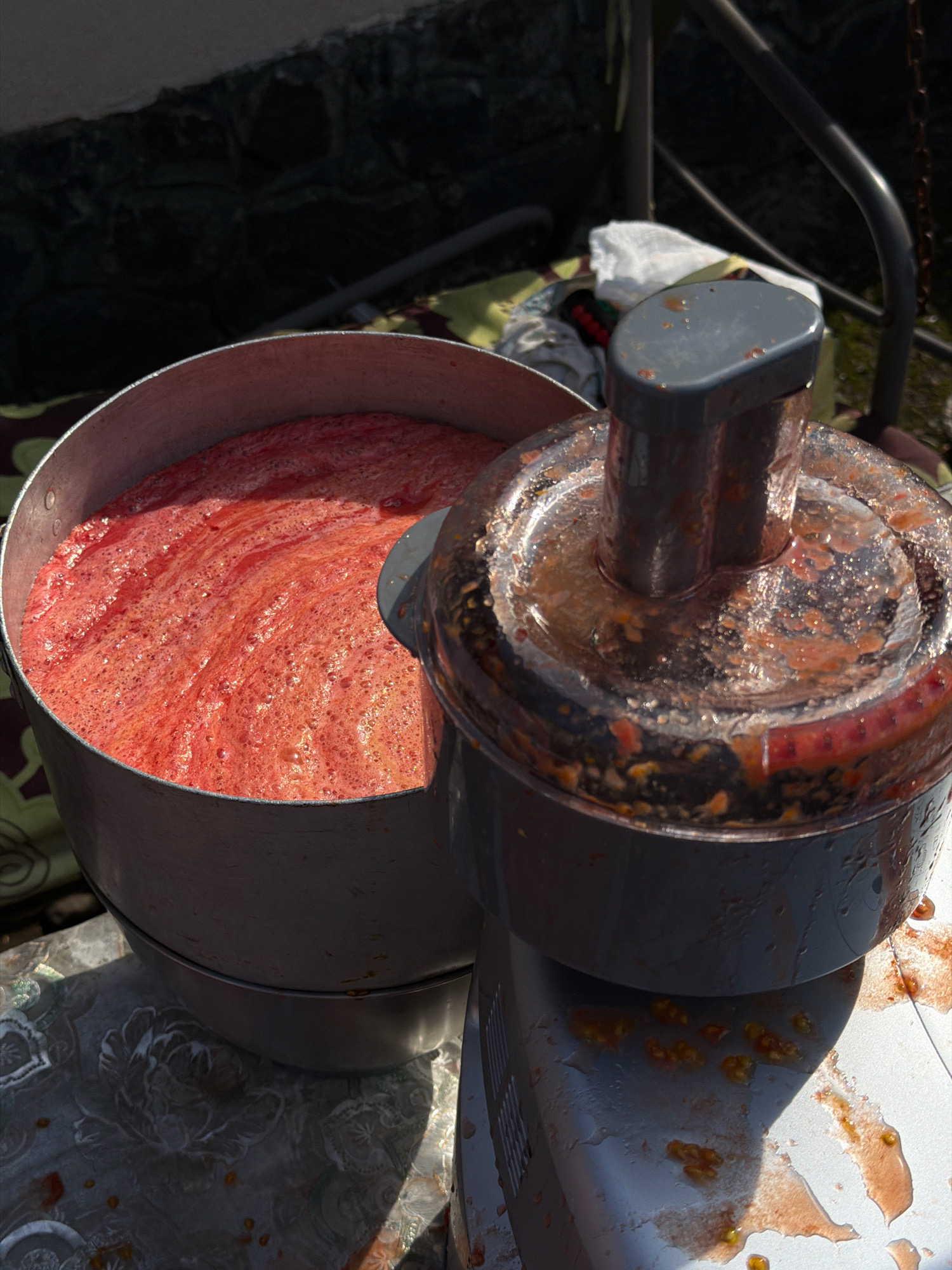 In Novosilka, Ukraine, a close-up, high-angle shot captures the aftermath of processing tomatoes. A large, silver pot overflows with a frothy, red liquid, likely tomato puree, while a tomato processor with splatters of the same red substance sits to the right. The scene appears to be outdoors, with a hint of dark stonework and greenery in the background, suggesting a residential yard or garden. A patterned cloth and a white fabric are visible near the processor, possibly used for cleanup or as a protective layer. The sun casts strong shadows, indicating a bright day. The overall impression is one of domestic food preparation, likely for canning or making sauce.