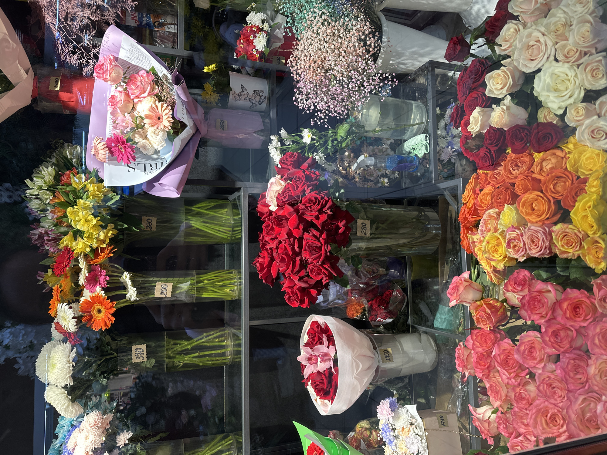The image depicts a storefront flower shop in Chaikovsky, Russia. Various floral arrangements and individual flower types are displayed behind a glass window. Roses in shades of red, white, yellow, orange, and pink are densely grouped. Additional flower varieties are arranged in vases or bouquets, including Gerbera daisies, chrysanthemums, and baby's breath. Price tags are visible on the arrangements.