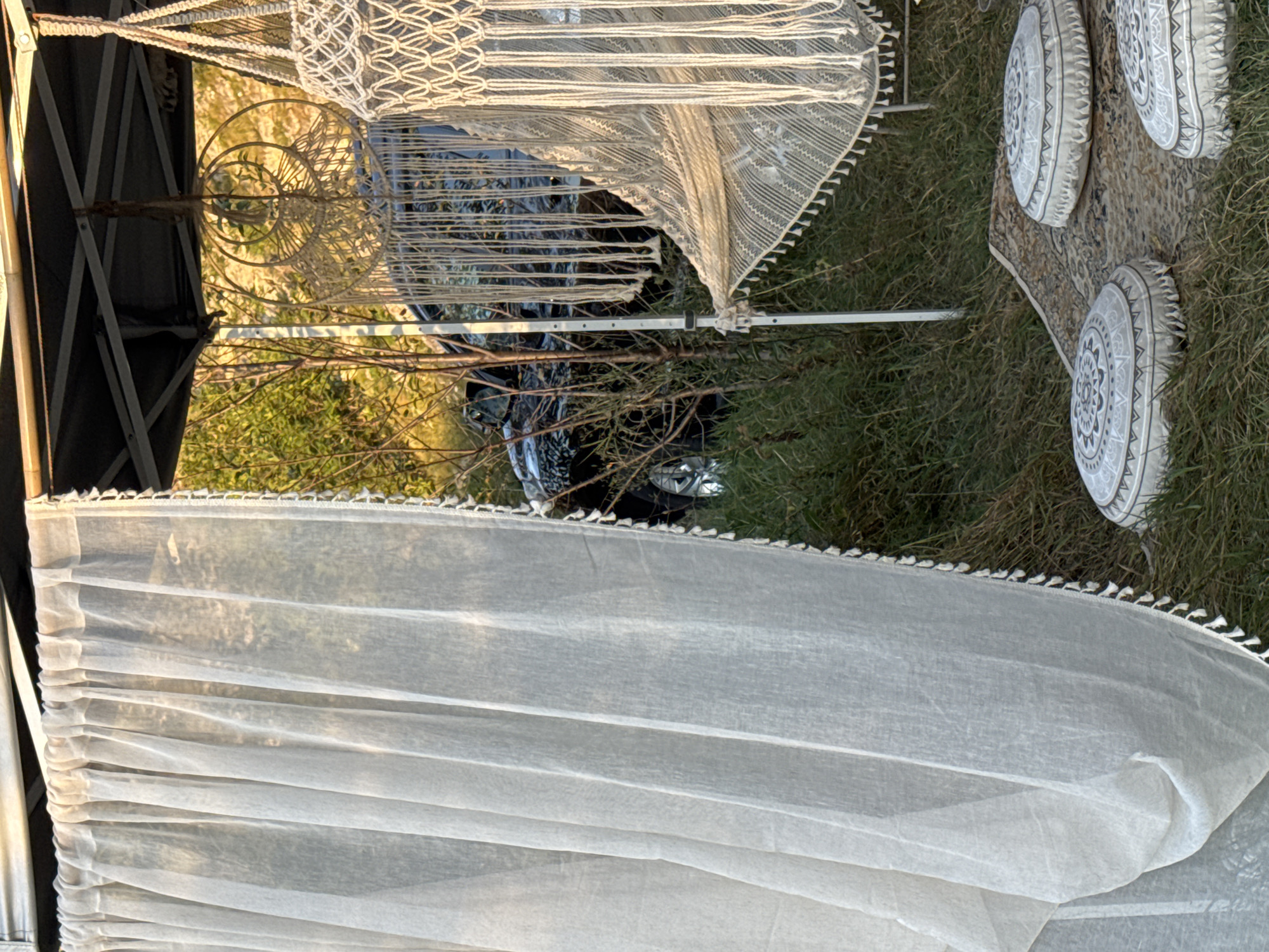 The image depicts an outdoor scene in Trebujeni, Moldova, characterized by a bohemian-style setup. In the foreground, a sheer white curtain with small pom-poms along the edge hangs to the left, allowing a glimpse of greenery and a vehicle behind it. To the right, a macrame hammock is suspended from a canopy structure, with another macrame decoration hanging above it. Three round floor cushions with intricate grey patterns rest on a patterned rug, which is placed on the grassy ground. In the background, a dark-colored vehicle is partially visible amidst the trees and vegetation. The overall impression is that of a cozy, decorated outdoor space, possibly for relaxation or camping.