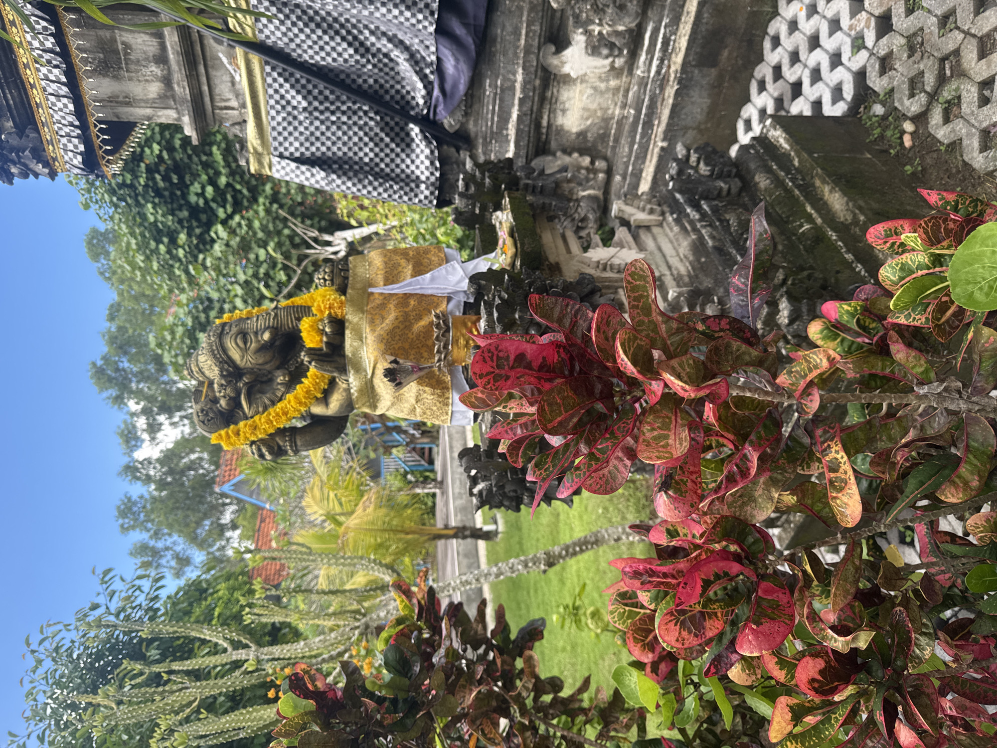 The image depicts a statue of an elephant-headed deity, likely Ganesha, adorned with a yellow garland and clothed in a gold and white fabric, positioned within a garden setting in Nusa Penida, Indonesia. The statue stands in front of a stone structure draped with a checkered cloth. The foreground is filled with red and green Croton plants. In the background, there is a glimpse of a blue and white structure and tropical foliage. The sky is visible in the upper part of the image.