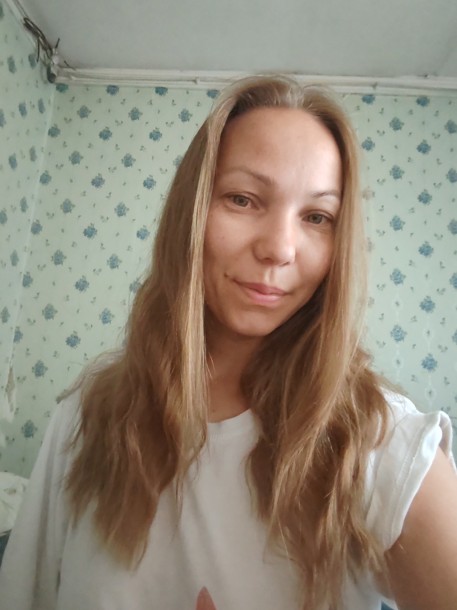 A woman is taking a selfie indoors. She has long, wavy, light brown hair and is wearing a white t-shirt. She is looking directly at the camera with a slight smile. The background is a wall with light green floral wallpaper. There is some visible clutter on the wall near the ceiling, including wires. The image appears to be taken in a residential setting in the city of Chukhlem, Russia, as indicated by the context provided.