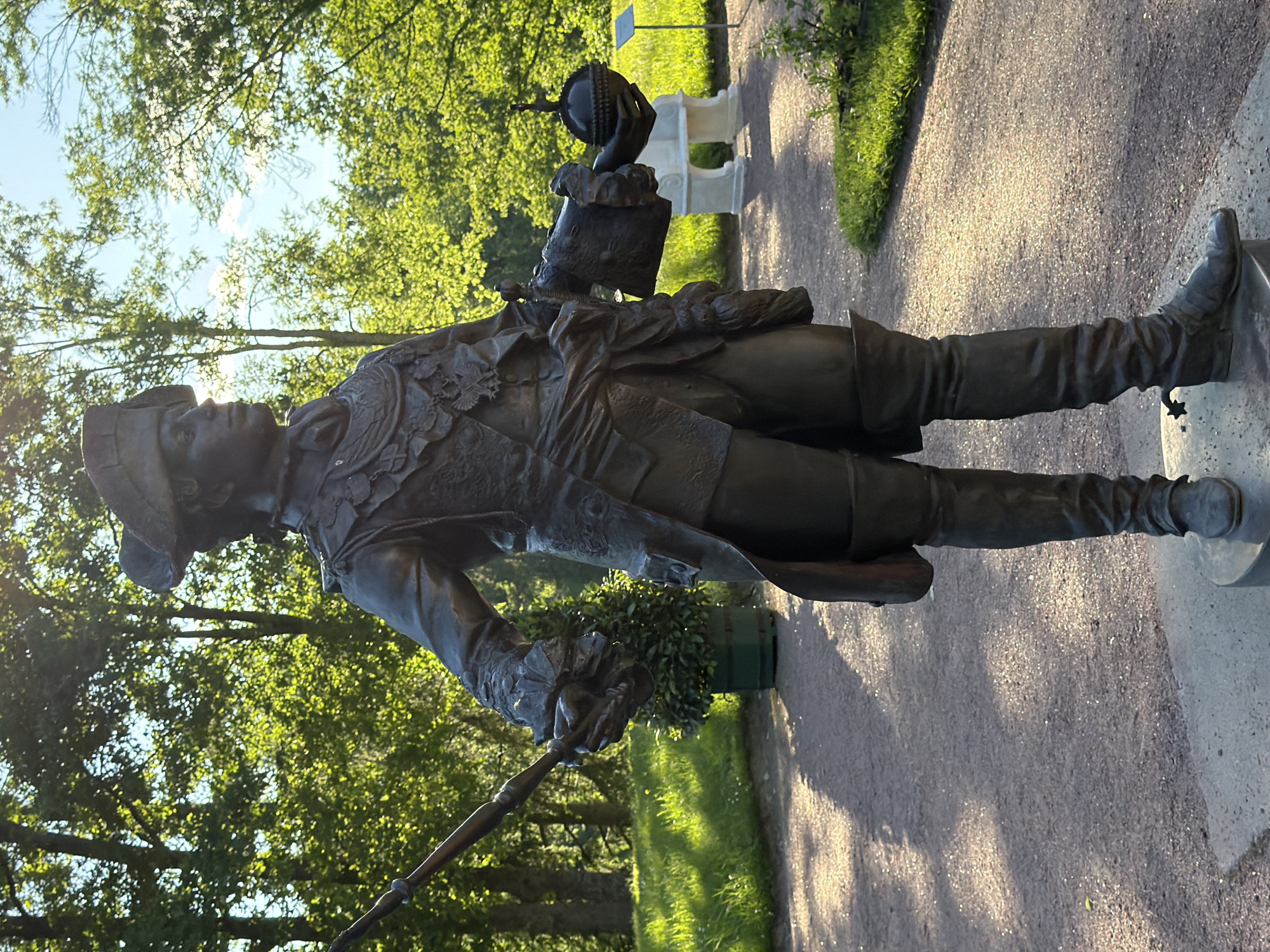 A full-body bronze statue of a man in historical military attire stands in a park on a sunny day. The statue is dressed in a wide-brimmed hat, a long coat with ornate trim, breeches, and knee-high boots. He holds a long, thin object in his left hand and a scepter or orb in his right. The statue is positioned on a circular stone base. Behind the statue, a pathway is visible, lined with trees and green grass. Sunlight filters through the leaves of the trees, creating dappled light on the ground. A potted plant is located to the left of the statue, and in the background, a white stone bench and a small sign are present. The sky is blue with some scattered clouds.
