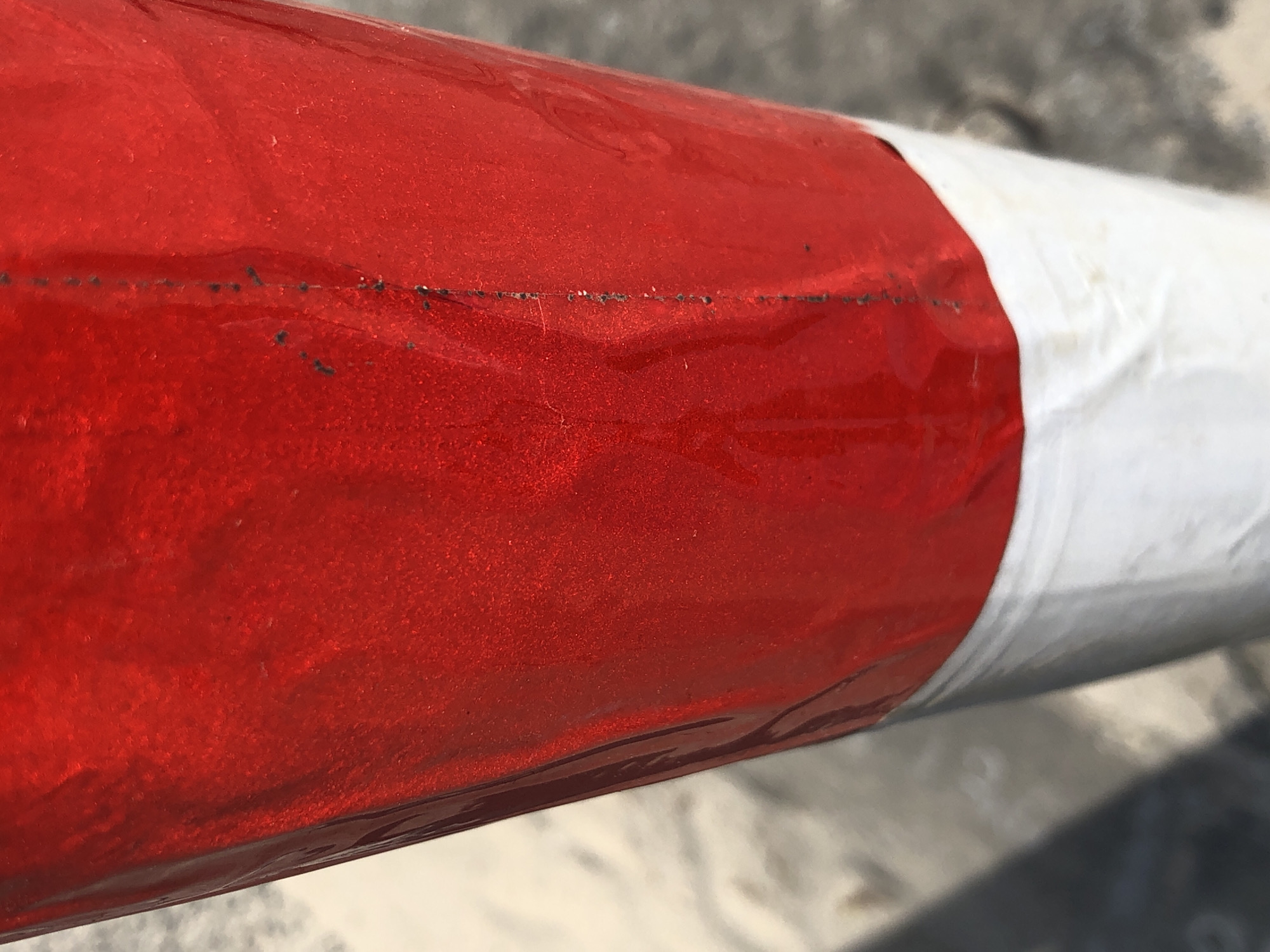 This media file is a close-up, eye-level shot of a cylindrical object wrapped in red and white tape, likely a buoy or marker. The object is positioned horizontally, with the red section taking up the majority of the frame on the left, and a white section visible on the right. The red tape appears to be a glossy, reflective material with small specks or imperfections visible along a horizontal line. The white tape looks more matte and slightly textured. The background is blurred, showing hints of a textured surface, possibly concrete or asphalt, and a lighter, out-of-focus area that could be sand or water. The lighting suggests daylight, but the time of day and weather are not clearly discernible. There is no visible text in the image. The context of Da Nang, Vietnam, might suggest the object is part of a marine or coastal marker, possibly near a beach or waterway, used for navigation or safety. The focus is entirely on the texture and detail of the object's surface.